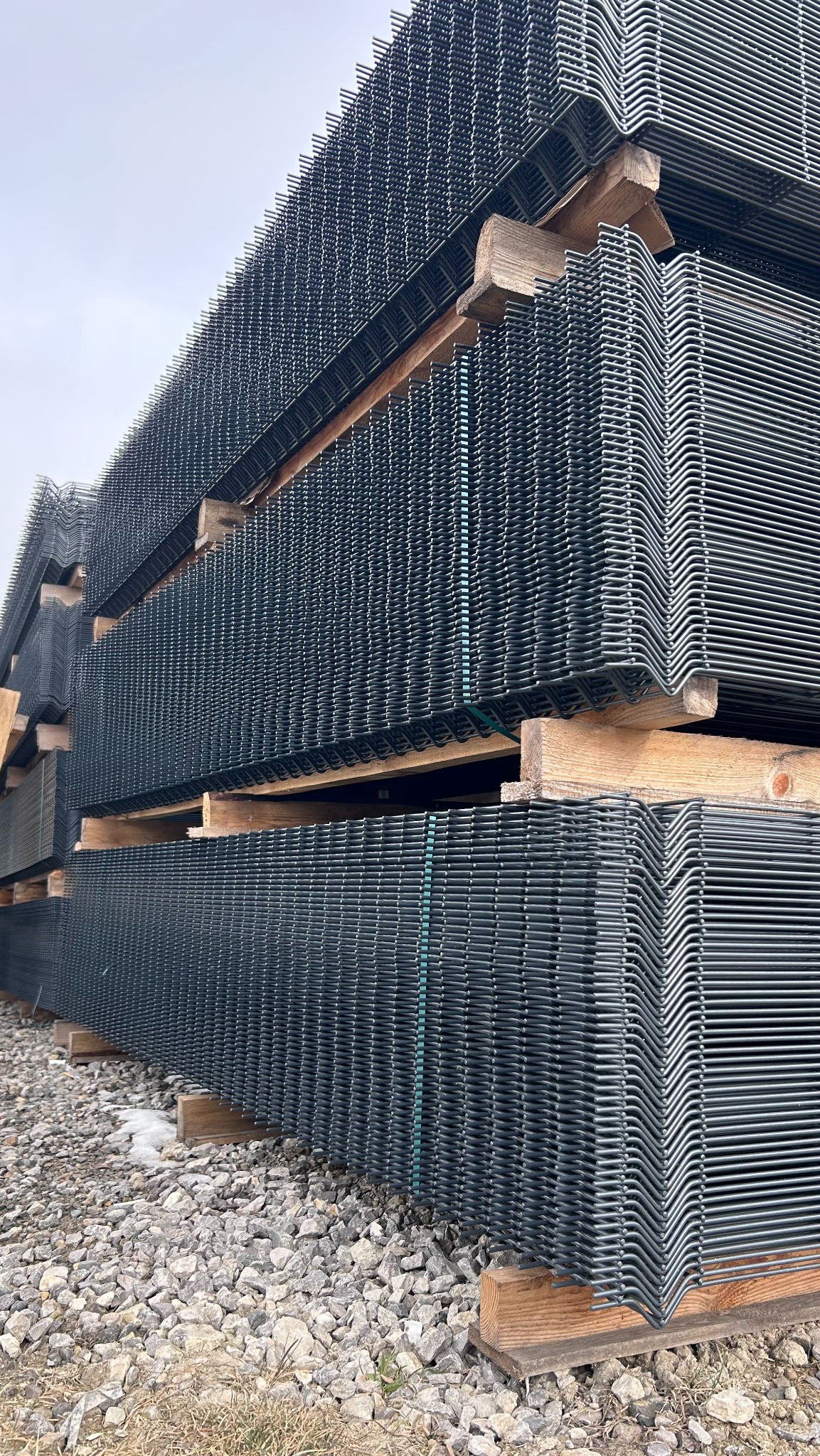 Stos paneli ogrodzeniowych zgrzewanych, ułożonych na drewnianych paletach na żwirowym podłożu, gotowych do transportu i montażu.