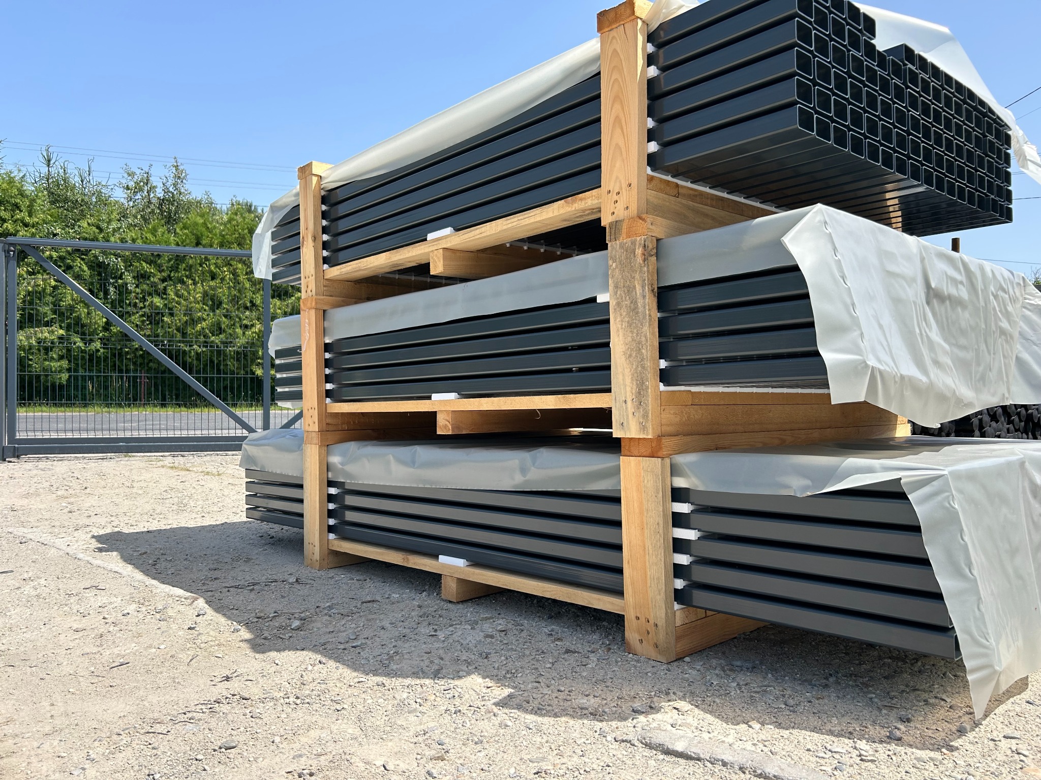 Stos paneli ogrodzeniowych w drewnianej ramie, zabezpieczonych folią, na żwirowym podłożu. W tle widoczne zielone drzewa i fragment metalowej bramy.