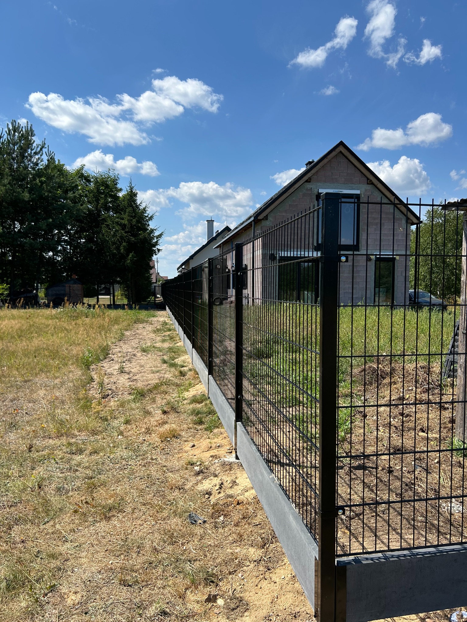 Nowoczesne ogrodzenie panelowe z podmurówką betonową, widok z perspektywy ścieżki wzdłuż ogrodzenia, dom w tle, błękitne niebo z chmurami.