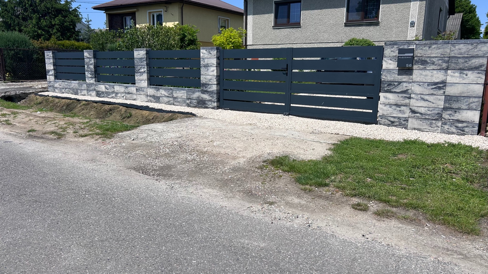 Nowoczesne, poziome przęsła z metalu w kolorze grafitowym, osadzone na słupkach z szarej kostki. Brama wjazdowa z tego samego materiału. Adres na słupku.