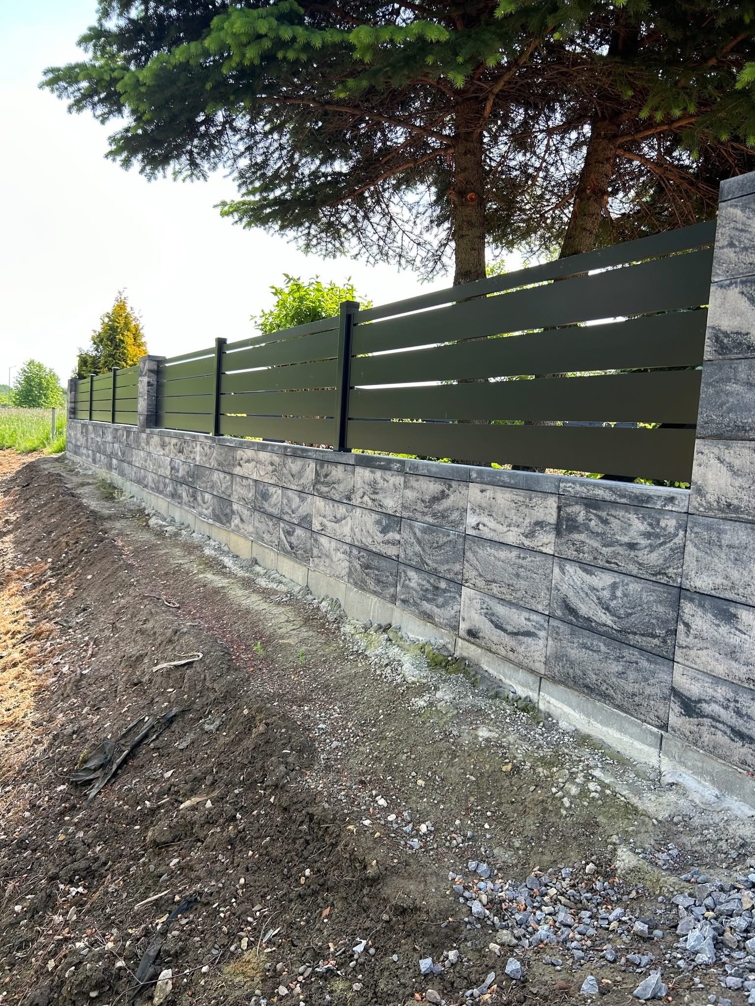 Nowoczesne ogrodzenie: poziome, zielone panele na murku z szarej kostki. W tle drzewa iglaste i trawa. Widok z poziomu gruntu, częściowo ukończony teren.