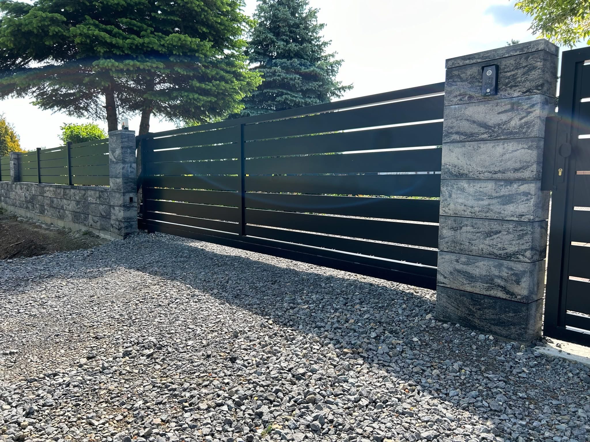 Nowoczesna, przesuwna brama z poziomych profili, zamontowana między kamiennymi słupkami z domofonem. Podłoże wysypane grysem, w tle drzewa.