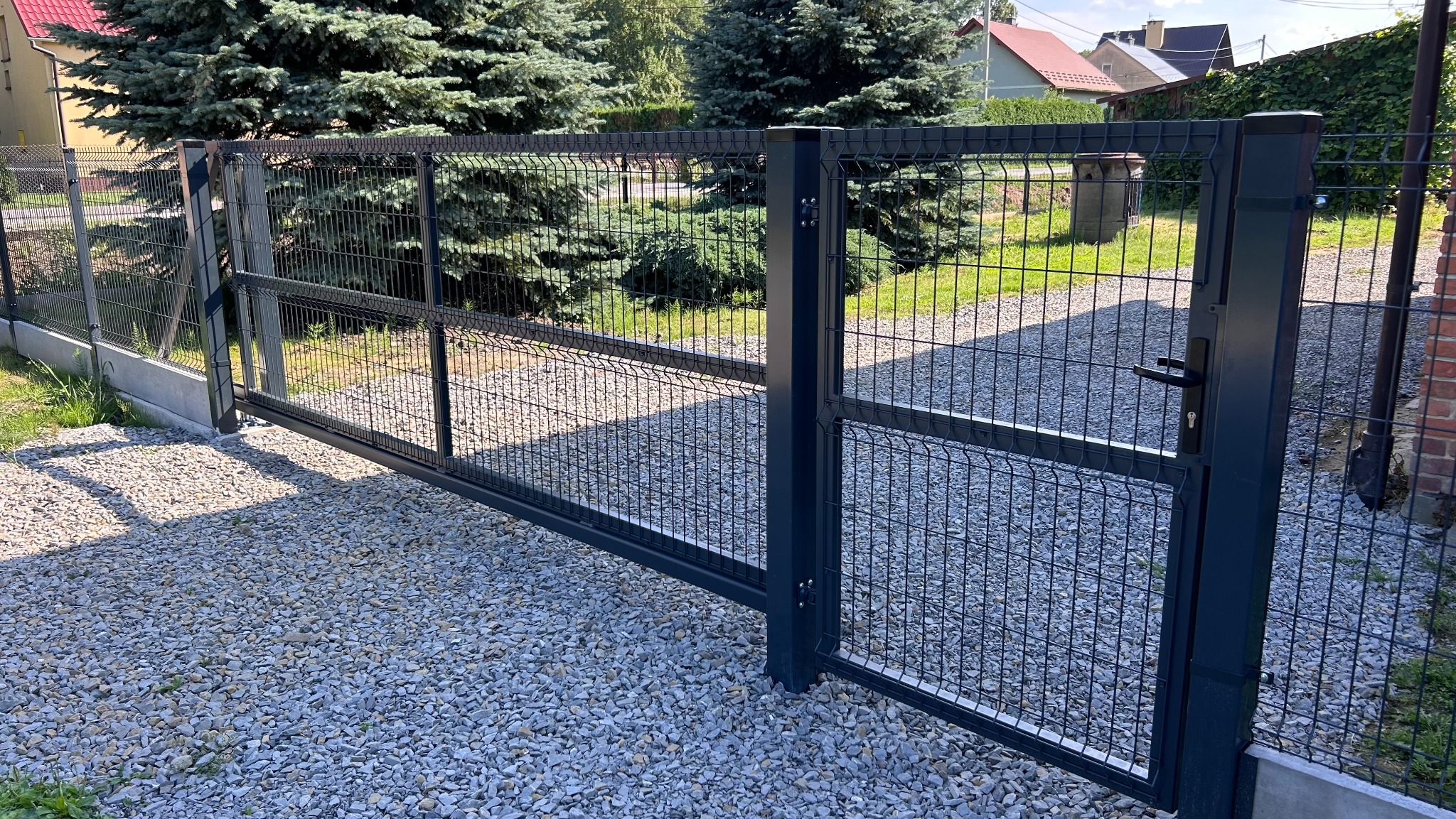 Grafitowa brama wjazdowa z siatki panelowej na żwirowym podłożu, z widocznym zamkiem i klamką, na tle zieleni i zabudowań.