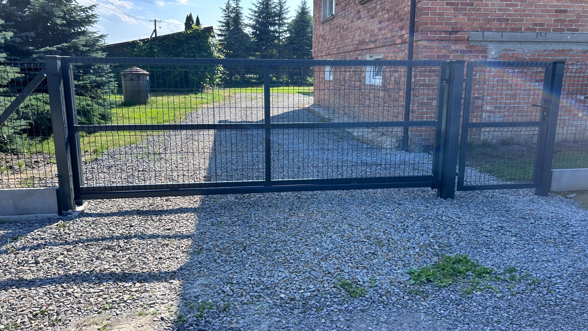 Nowoczesna, grafitowa brama z siatki z metalową ramą, na tle żwirowego podjazdu i ceglanego budynku. Widoczne drzewa iglaste w tle.