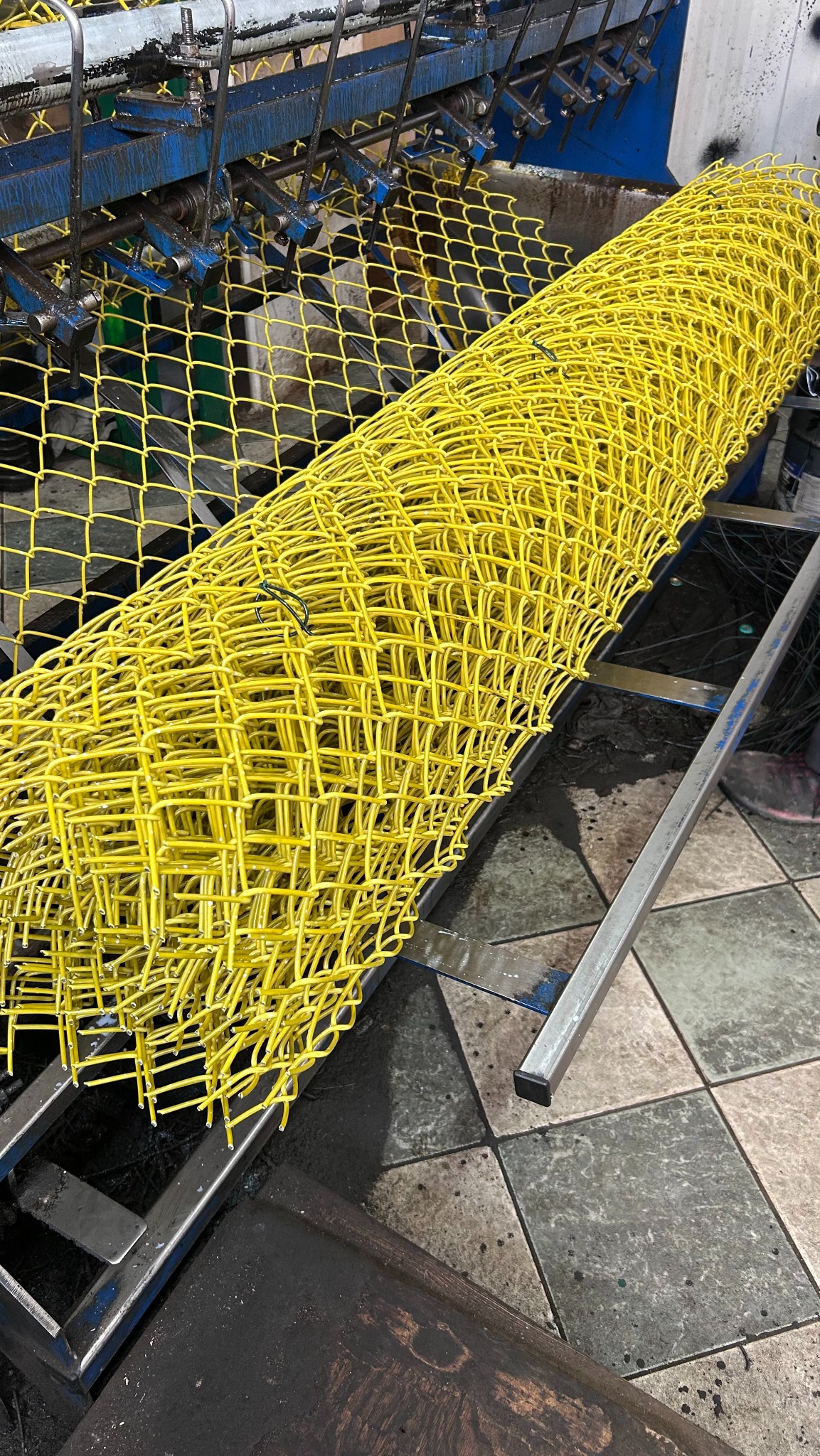 Żółta siatka ogrodzeniowa zwinięta w rolkę na metalowej konstrukcji maszyny produkcyjnej. Widoczne detale łączeń i faktura siatki.