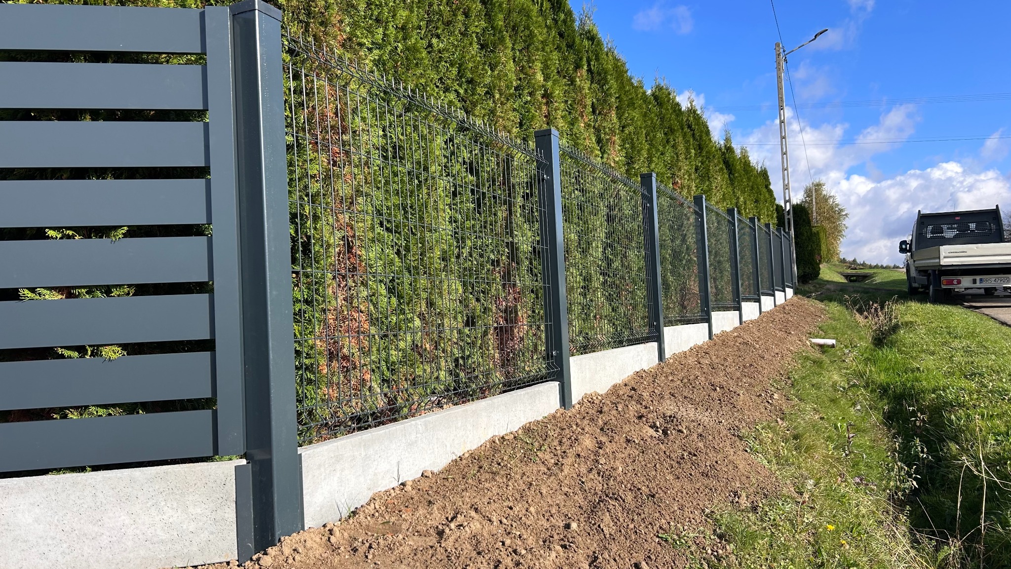 Nowoczesne ogrodzenie: szare panele z poziomymi szczelinami i siatka na betonowej podmurówce, tło z zielonych tuj, błękitne niebo. Widok z boku, lekki skos.