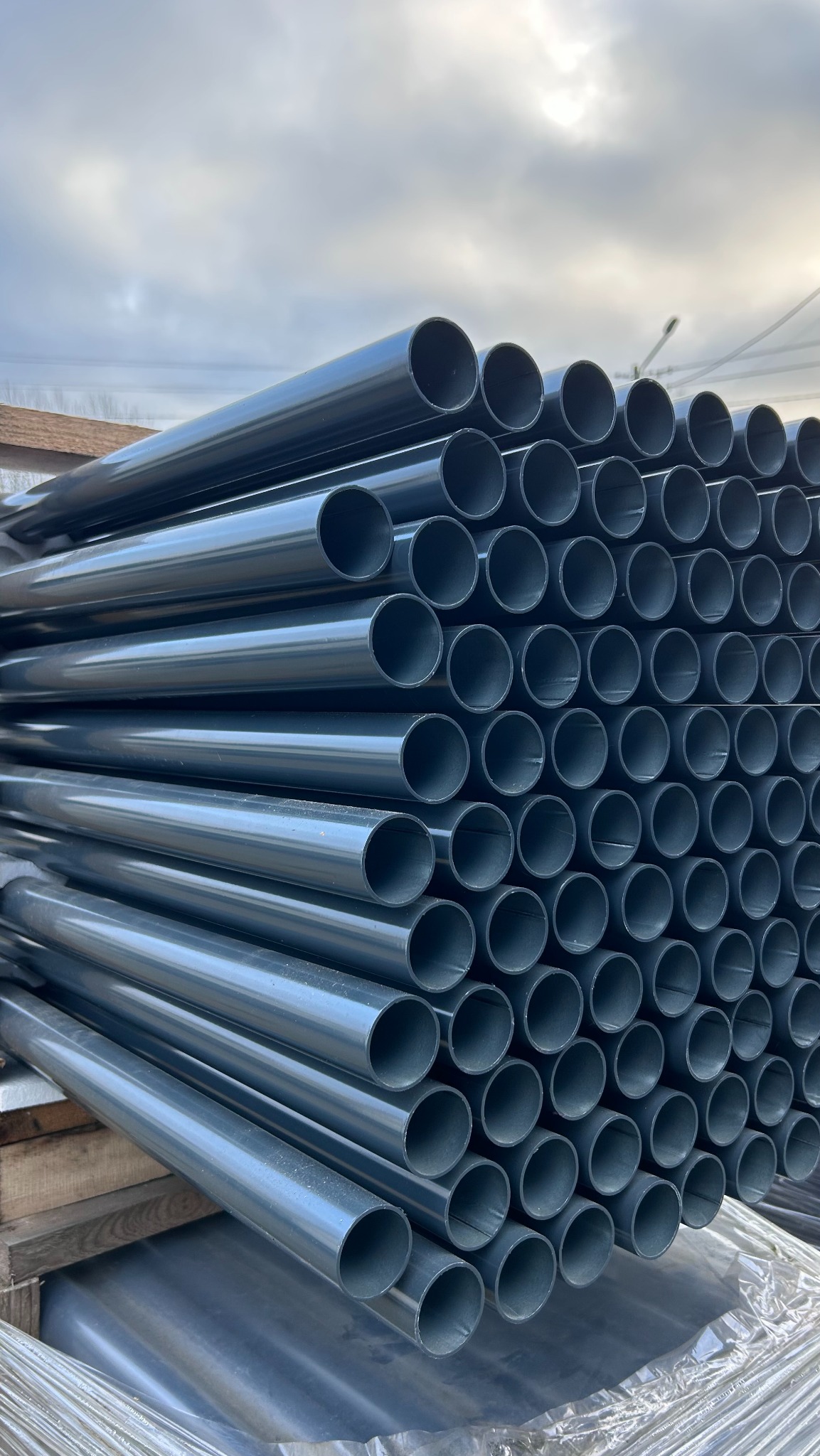 Stos rur metalowych w kolorze grafitowym, ułożonych równolegle na drewnianej palecie, z widocznym niebem w tle. Elementy do konstrukcji.