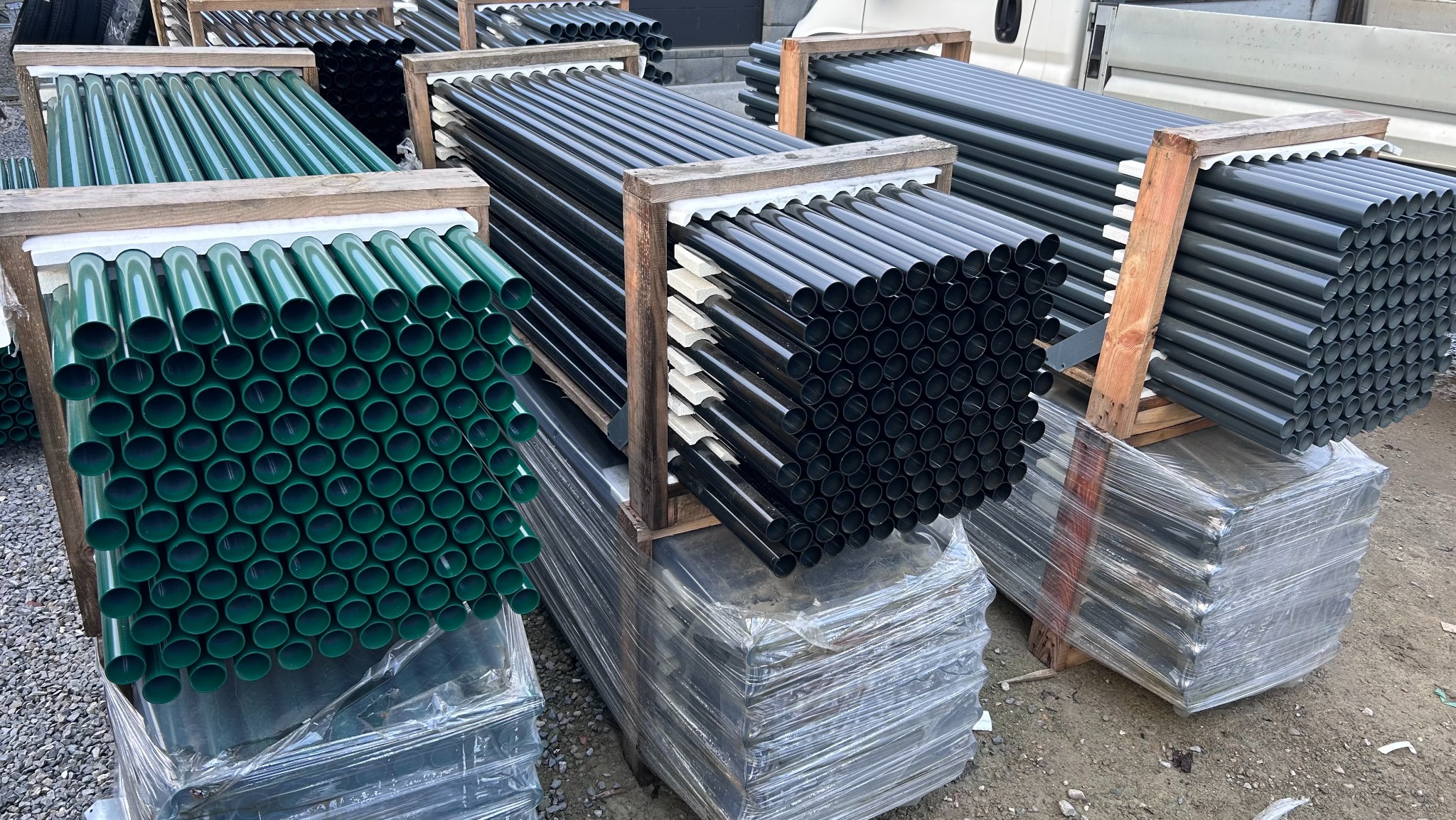 Pakiety rur do ogrodzeń w odcieniach zieleni, czerni i szarości, ułożone w drewnianych ramach i owinięte folią, gotowe do transportu. Widok z bliska.