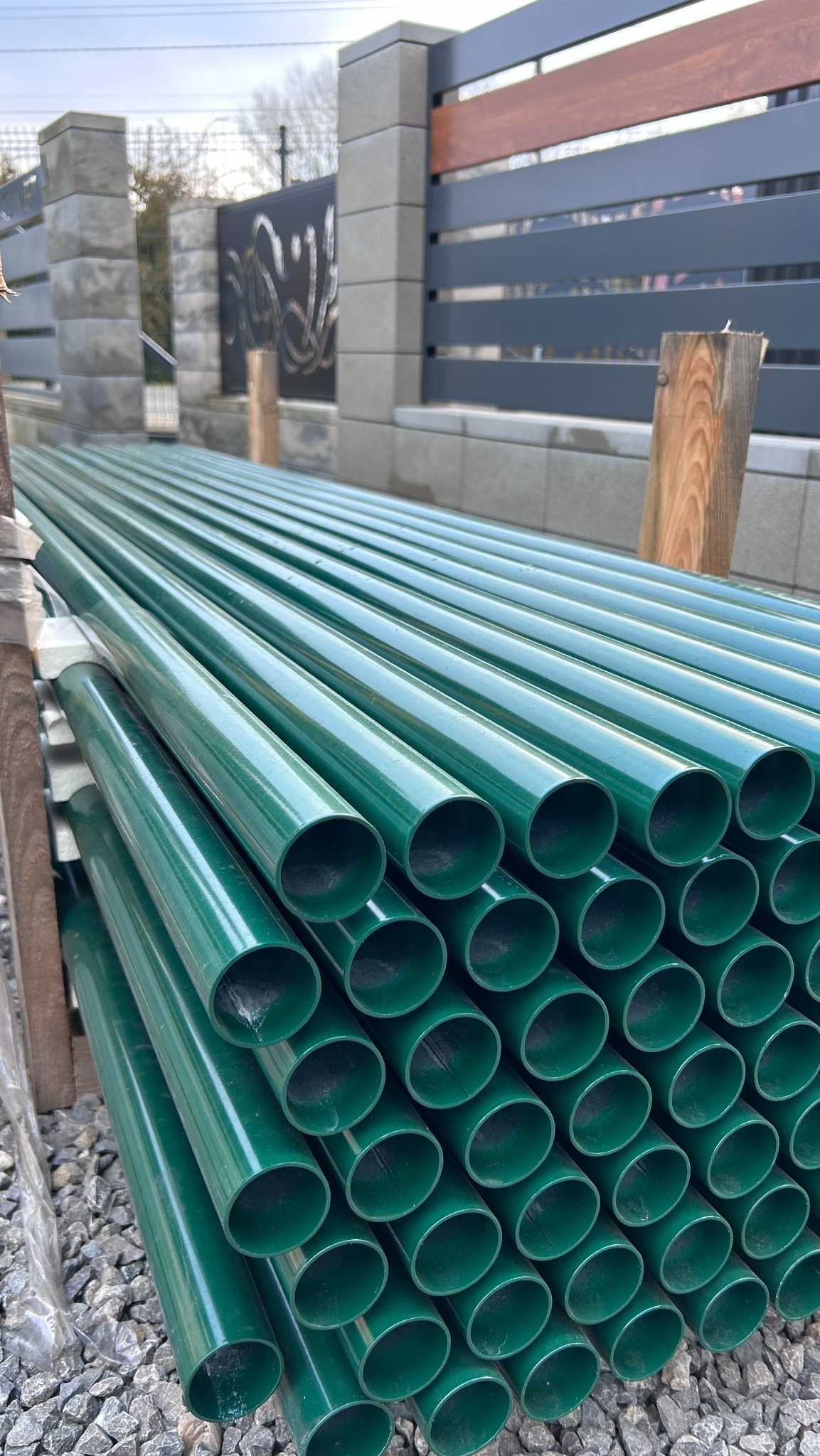 Stos zielonych, metalowych słupków ogrodzeniowych, ułożonych warstwami na drewnianych belkach, z fragmentem nowoczesnego ogrodzenia w tle.