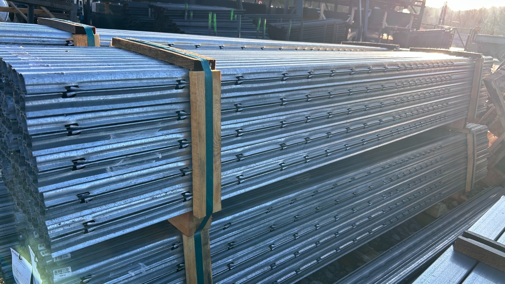 Stos paneli ogrodzeniowych z metalu, zabezpieczonych drewnianymi belkami i taśmą, gotowych do montażu. Widoczne mocowania paneli. Słoneczne oświetlenie.