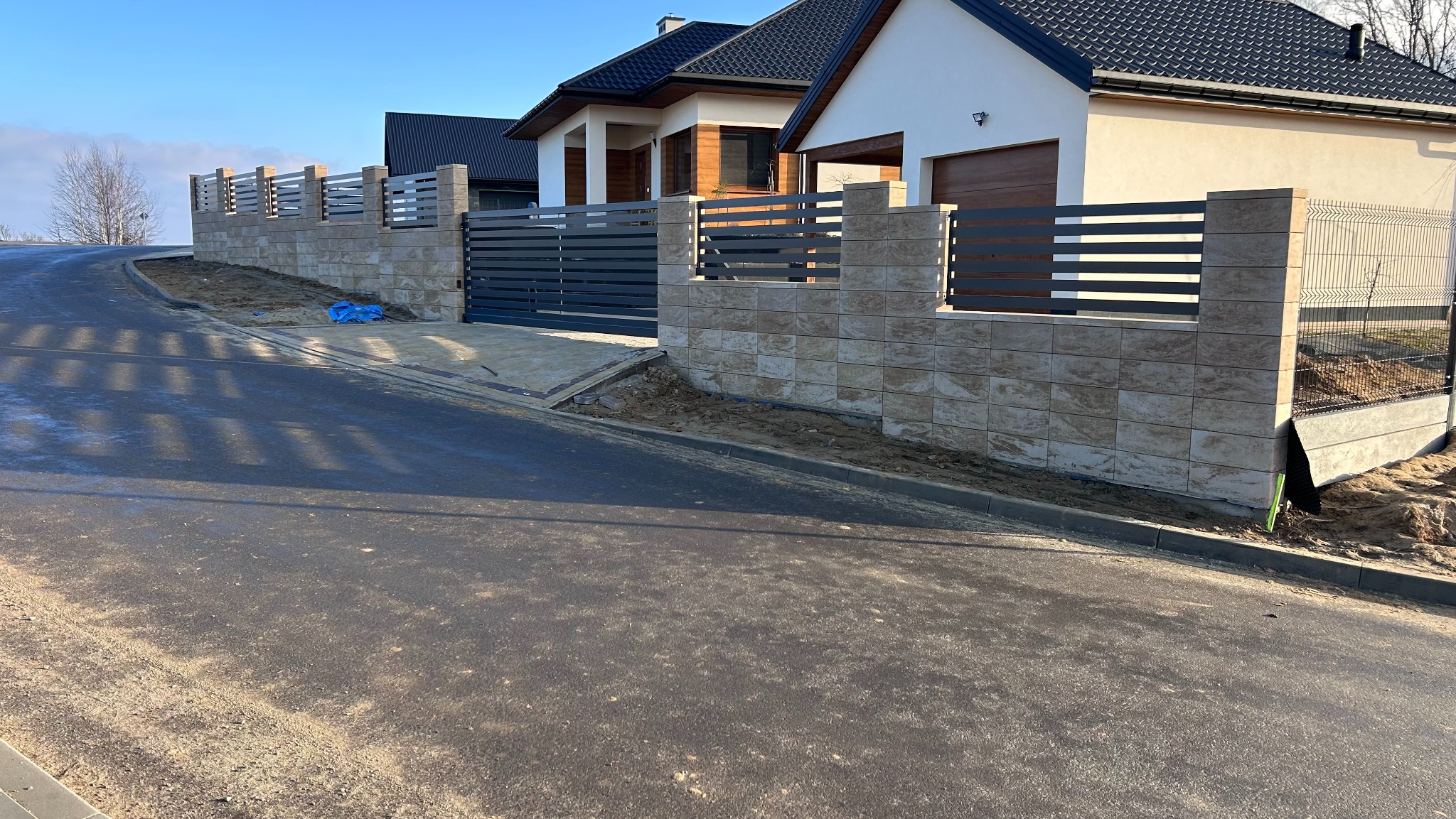 Nowoczesne ogrodzenie z metalowymi przęsłami i elementami z bloczków betonowych, z bramą wjazdową, na tle domu jednorodzinnego i asfaltowej drogi.