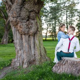 Niezapomniane wspomnienia - Para młoda w objęciach siedzi na pniu drzewa, panna młoda w wianku i jeansowej kurtce, pan młody w białej koszuli z czerwonymi szelkami, w tle ruiny budynku i drzewa.