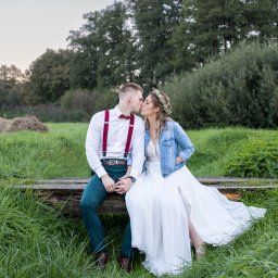 Niezapomniane wspomnienia - Para młoda, on w zielonych spodniach i bordowych szelkach, ona w białej sukni i jeansowej kurtce, całują się siedząc na drewnianej kłodzie na tle zieleni.