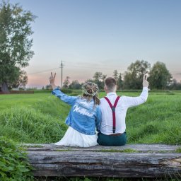 Niezapomniane wspomnienia - Para młoda siedzi tyłem na drewnianej kłodzie na tle zielonego pola i drzew, ona w wianku i kurtce jeansowej z napisem 'Just Married', oboje z podniesionymi rękami pokazują znak zwycięstwa.
