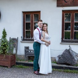 Niezapomniane wspomnienia - Uśmiechnięta para narzeczonych w objęciach na tle białej ściany z drewnianymi oknami i rustykalnymi dekoracjami.