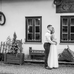 Niezapomniane wspomnienia - Czarno-białe zdjęcie pary młodej, przytulonej przed budynkiem z rustykalnymi elementami dekoracyjnymi, takimi jak drewniane okiennice, latarnia i napis 'CZARD'.