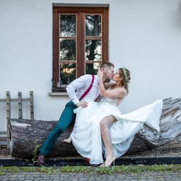 Niezapomniane wspomnienia - Pocałunek młodej pary siedzącej na spróchniałym pniu drzewa przed białym budynkiem z drewnianymi oknami, panna młoda z wiankiem na głowie i w białej sukni, pan młody w zielonych spodniach i...