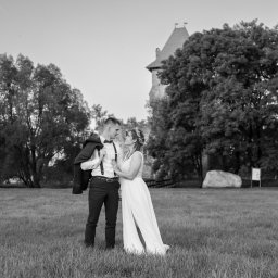 Niezapomniane wspomnienia - Czarno-białe zdjęcie narzeczonej w koronkowej sukni z rozcięciem i pana młodego z marynarką przewieszoną przez ramię, stojących na trawiastym polu z zamkiem w tle.