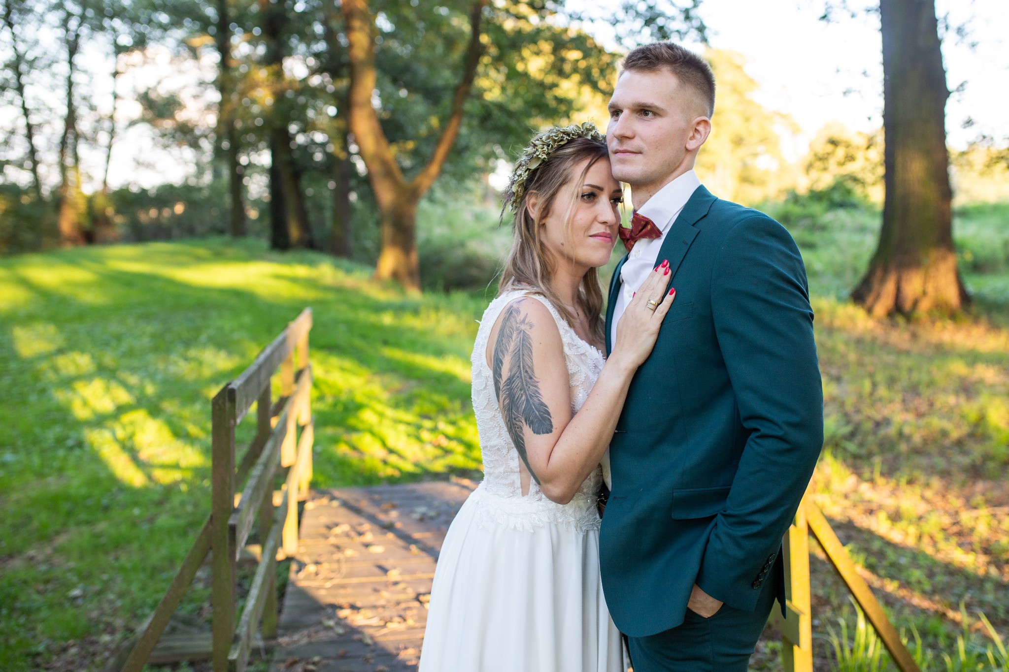 Para młoda w lesie, panna młoda w koronie z kwiatów i tatuażem pióra na ramieniu, pan młody w zielonym garniturze i bordowej muszce, pozują na tle drewnianego mostku.