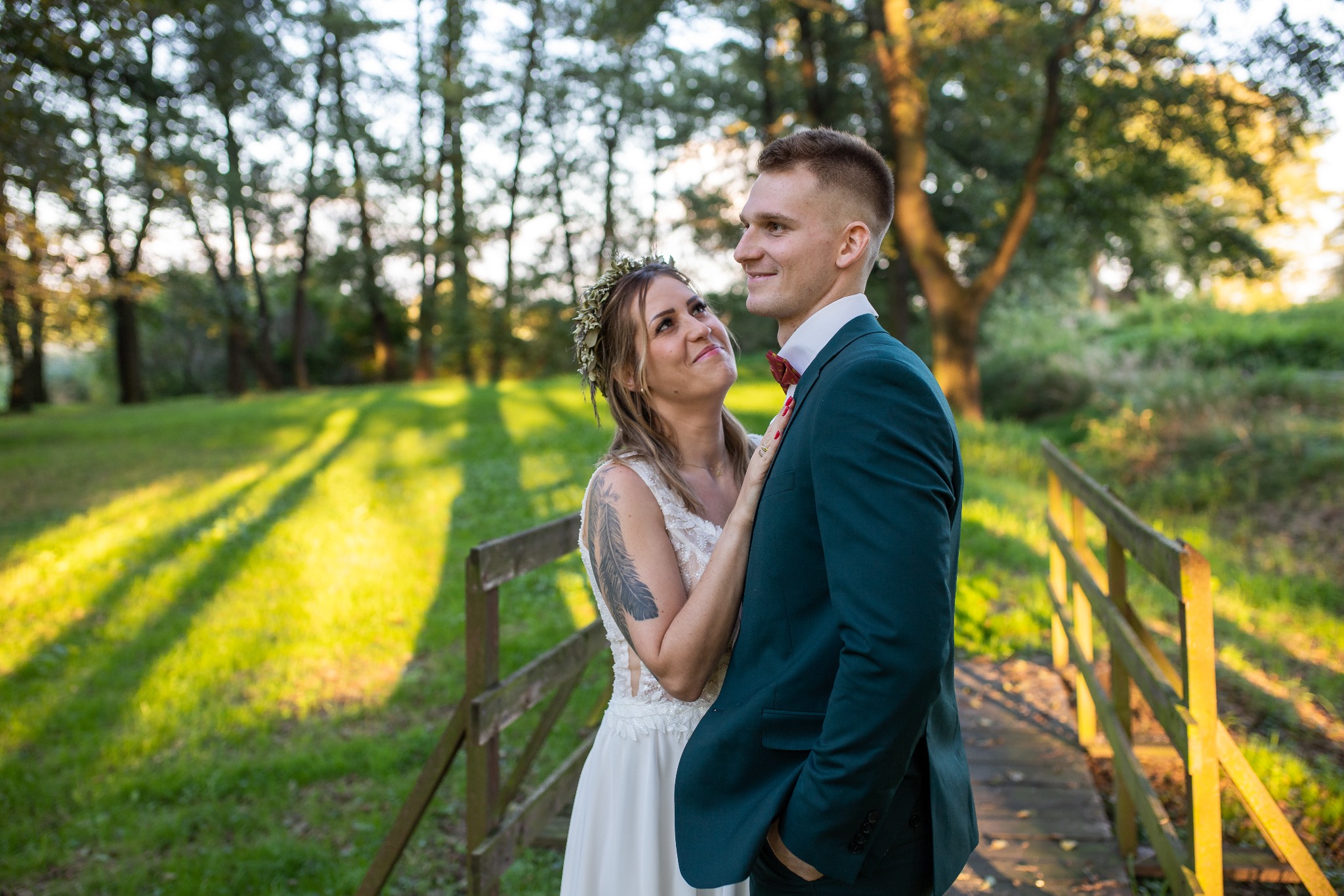 Para młoda na drewnianym mostku w lesie; panna młoda z wiankiem i tatuażem na ramieniu spogląda z miłością na pana młodego w ciemnozielonym garniturze.