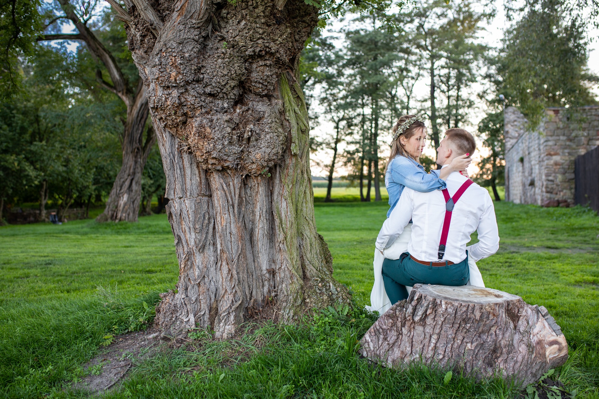 Para młoda w objęciach siedzi na pniu drzewa, panna młoda w wianku i jeansowej kurtce, pan młody w białej koszuli z czerwonymi szelkami, w tle ruiny budynku i drzewa.