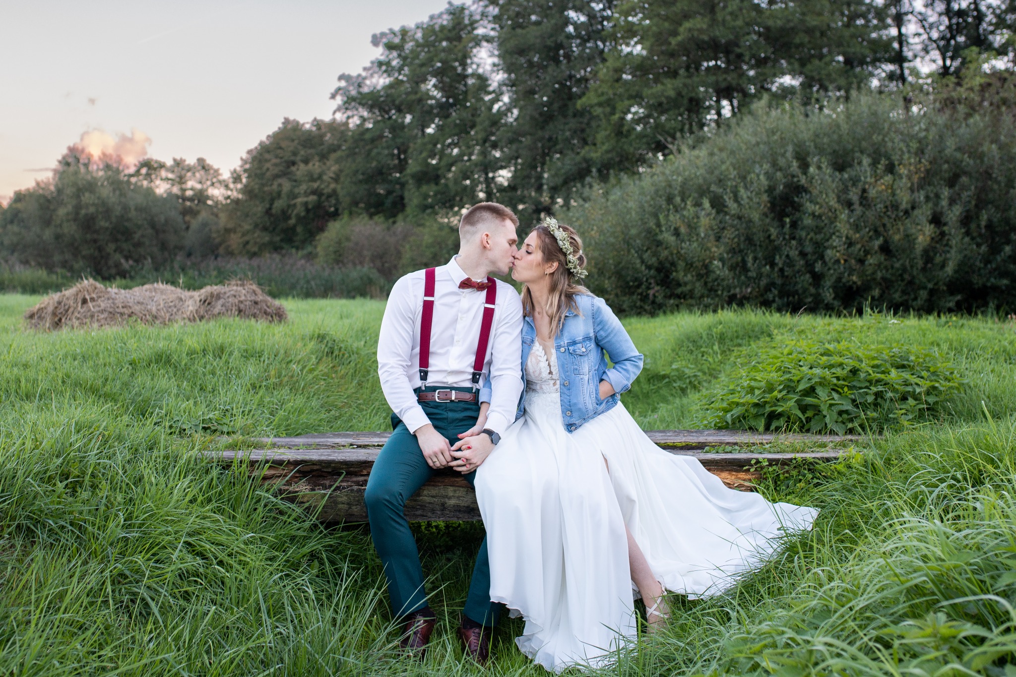 Para młoda, on w zielonych spodniach i bordowych szelkach, ona w białej sukni i jeansowej kurtce, całują się siedząc na drewnianej kłodzie na tle zieleni.