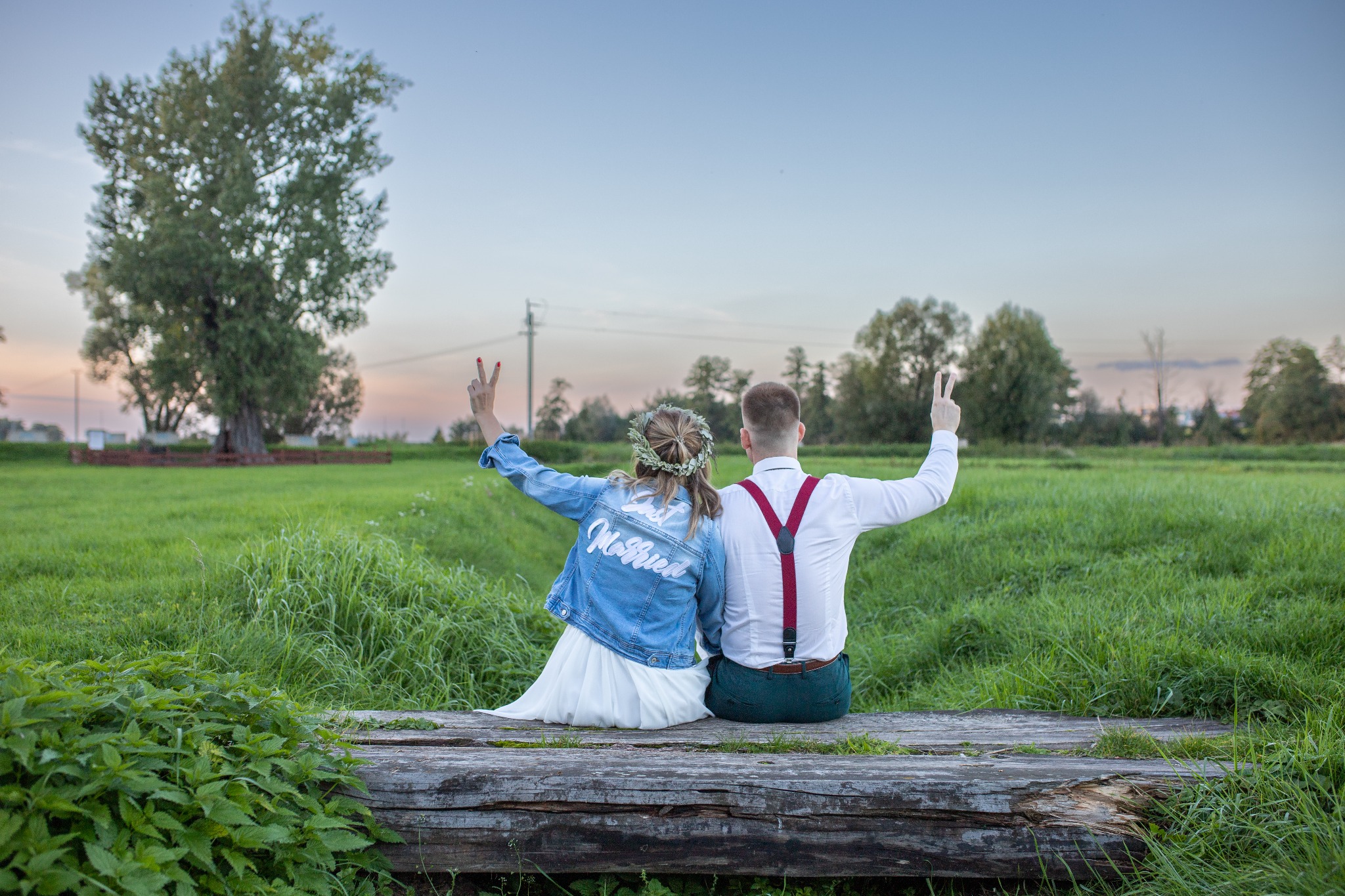 Para młoda siedzi tyłem na drewnianej kłodzie na tle zielonego pola i drzew, ona w wianku i kurtce jeansowej z napisem 'Just Married', oboje z podniesionymi rękami pokazują znak zwycięstwa.