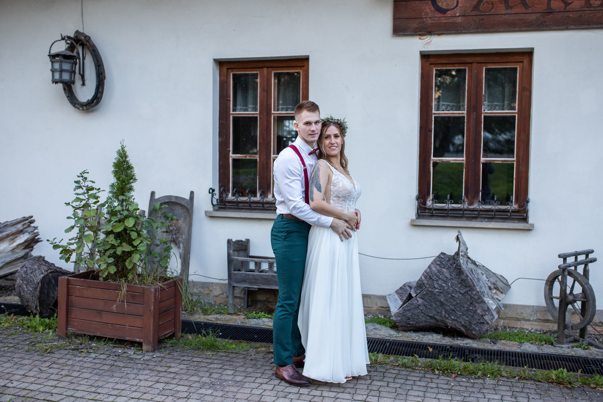 Uśmiechnięta para narzeczonych w objęciach na tle białej ściany z drewnianymi oknami i rustykalnymi dekoracjami.