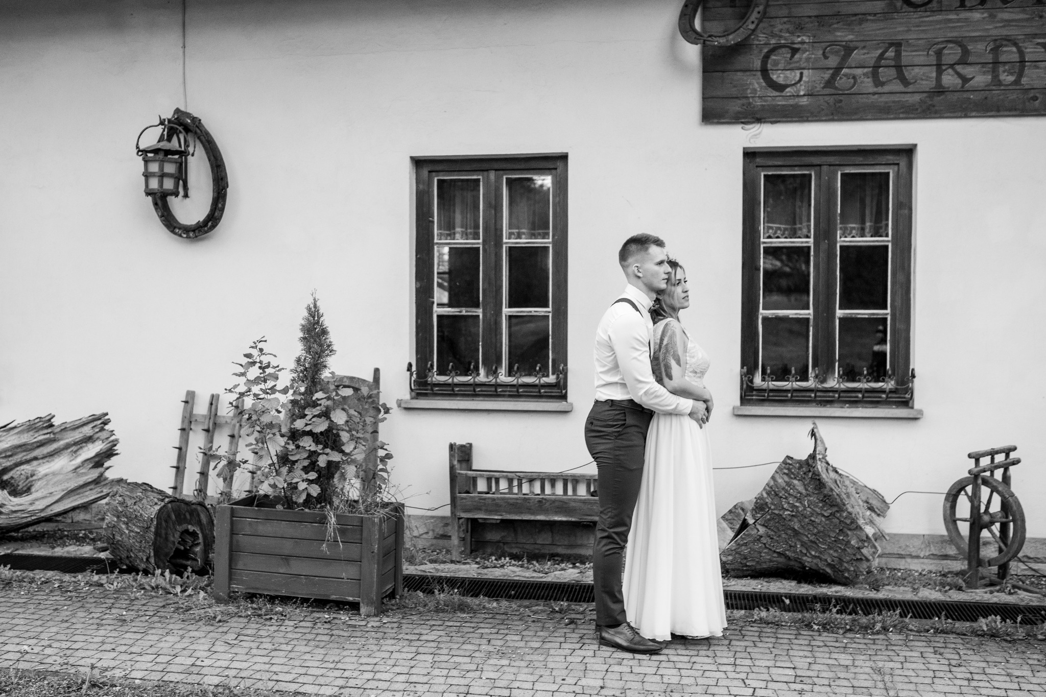 Czarno-białe zdjęcie pary młodej, przytulonej przed budynkiem z rustykalnymi elementami dekoracyjnymi, takimi jak drewniane okiennice, latarnia i napis 'CZARD'.