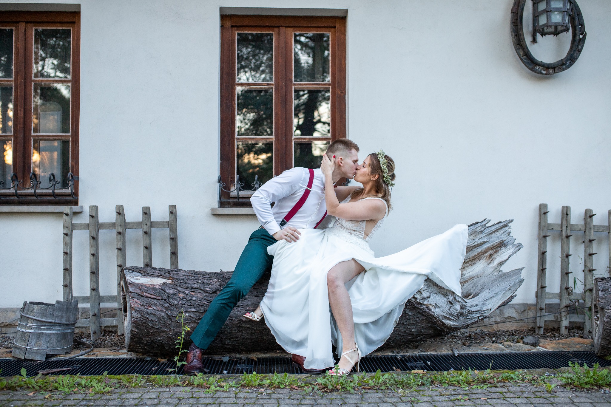 Pocałunek młodej pary siedzącej na spróchniałym pniu drzewa przed białym budynkiem z drewnianymi oknami, panna młoda z wiankiem na głowie i w białej sukni, pan młody w zielonych spodniach i...