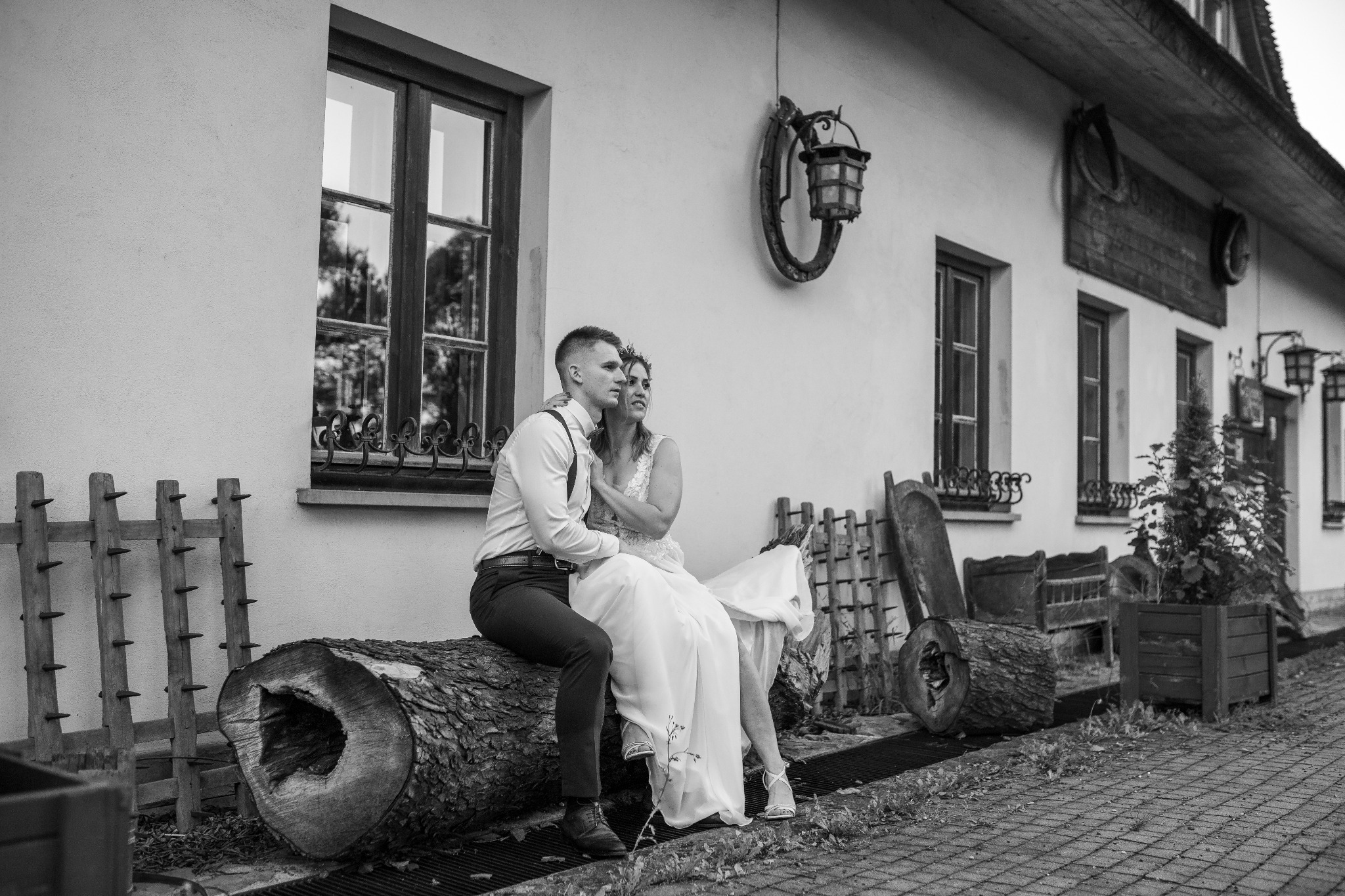 Czarno-białe zdjęcie pary młodej siedzącej na pniu drzewa przed budynkiem z oknami i ozdobnymi lampami, w tle widoczna drewniana konstrukcja z kolcami.