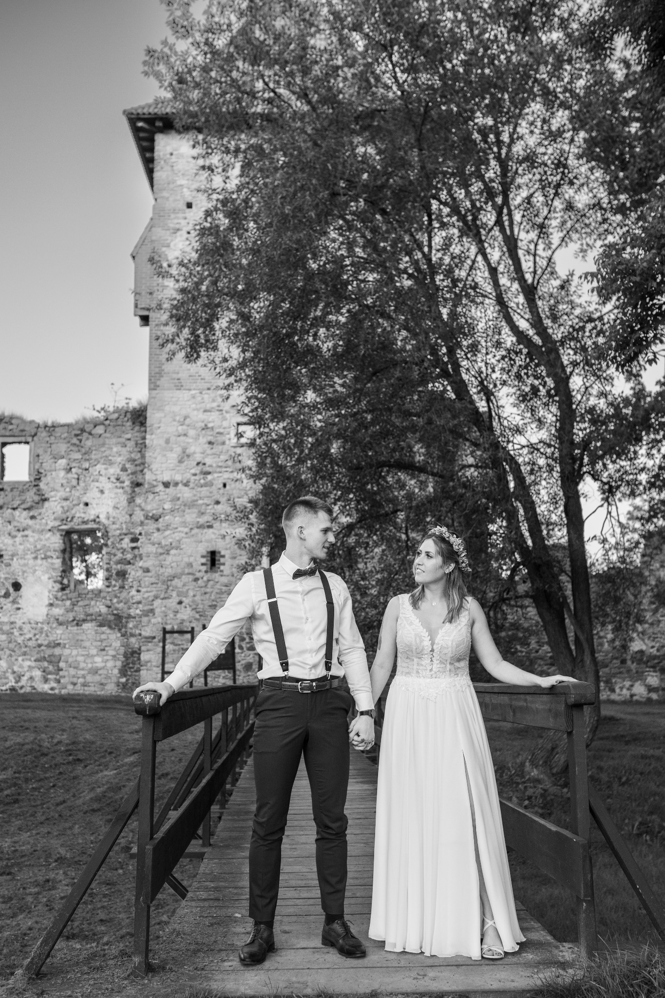 Czarno-białe zdjęcie pary młodej trzymającej się za ręce na drewnianym moście, w tle ruiny zamku i drzewo.