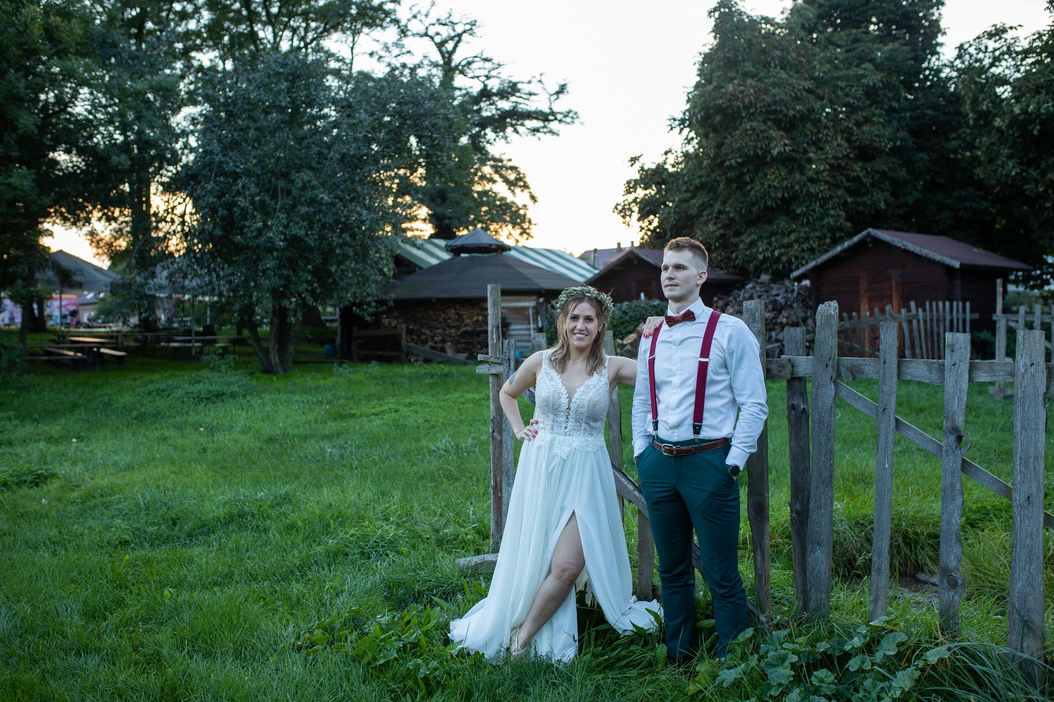Para młoda pozuje na tle drewnianego płotu i zieleni, ona w białej sukni z wiankiem, on w zielonych spodniach z szelkami i muszką.