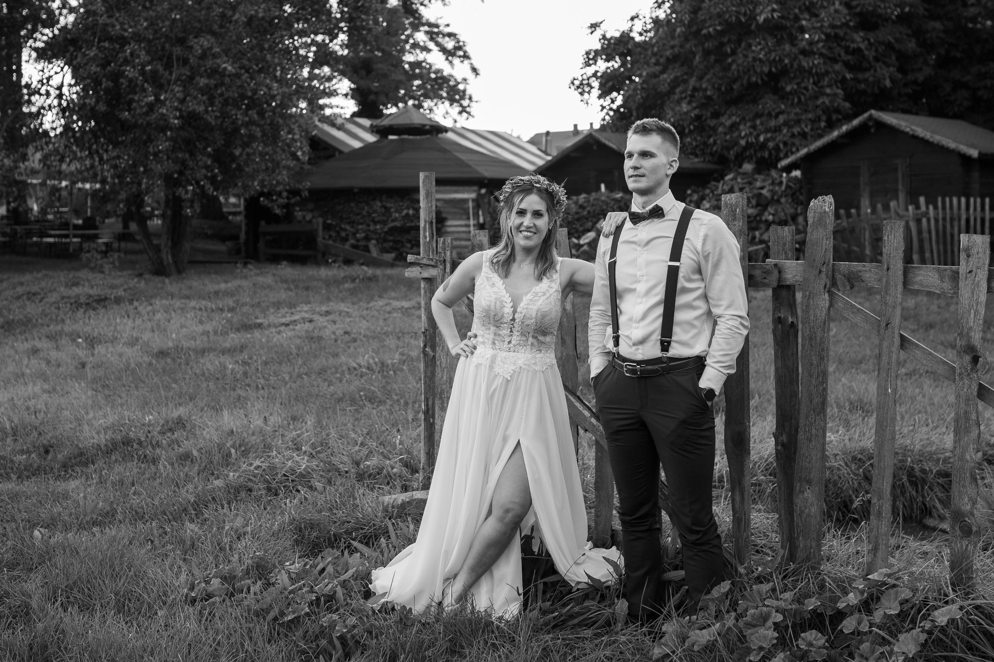 Czarno-białe zdjęcie pary młodej stojącej przy drewnianym płocie na tle wiejskich zabudowań; panna młoda w sukni z rozcięciem i wiankiem na głowie, pan młody w spodniach na szelkach i muszce.