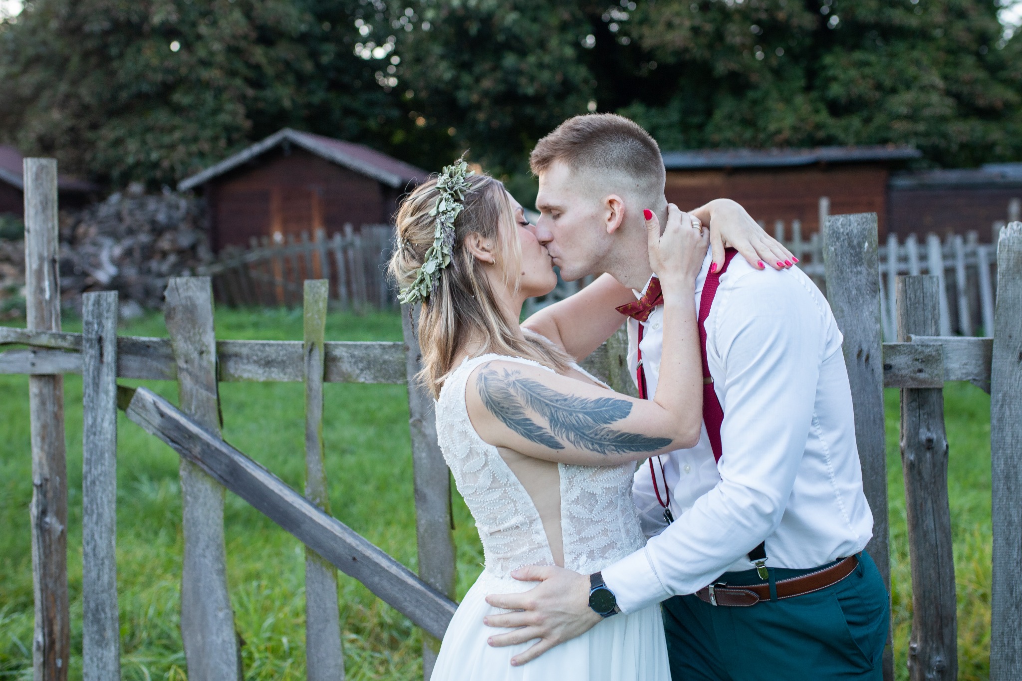 Para młoda całuje się na tle starego, drewnianego płotu i zieleni; panna młoda w sukni z koronkową górą i tatuażem na ramieniu, pan młody w białej koszuli, czerwonych szelkach i zielonych spodniach.