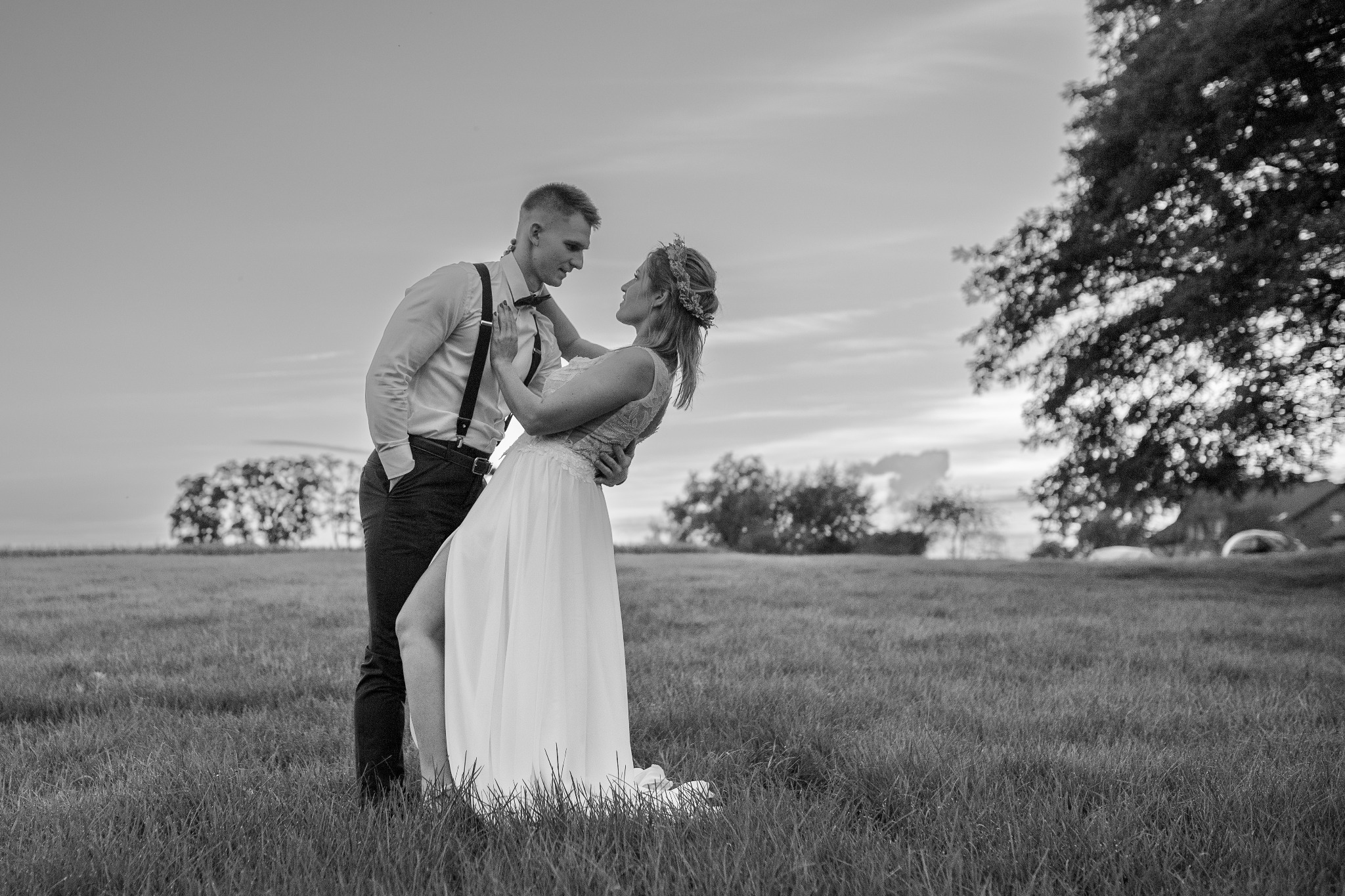 Czarno-białe zdjęcie pary młodej na łące; panna młoda w sukni z rozcięciem i wiankiem na głowie, pan młody w koszuli z szelkami, oboje patrzą na siebie z czułością.