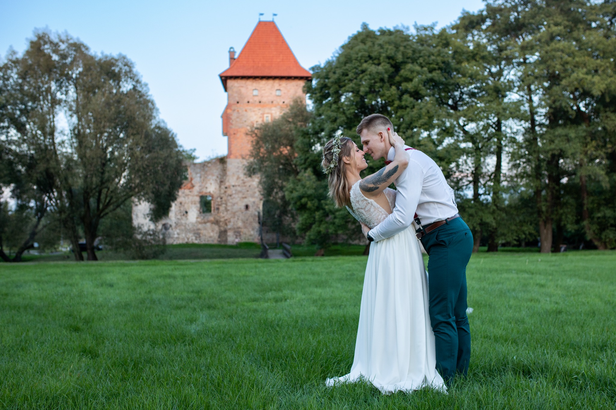 Para młoda przytulona na tle średniowiecznego zamku z czerwonym dachem, panna młoda w białej sukni z koronkowymi plecami i wiankiem, pan młody w białej koszuli i zielonych spodniach, oboje patrzą...