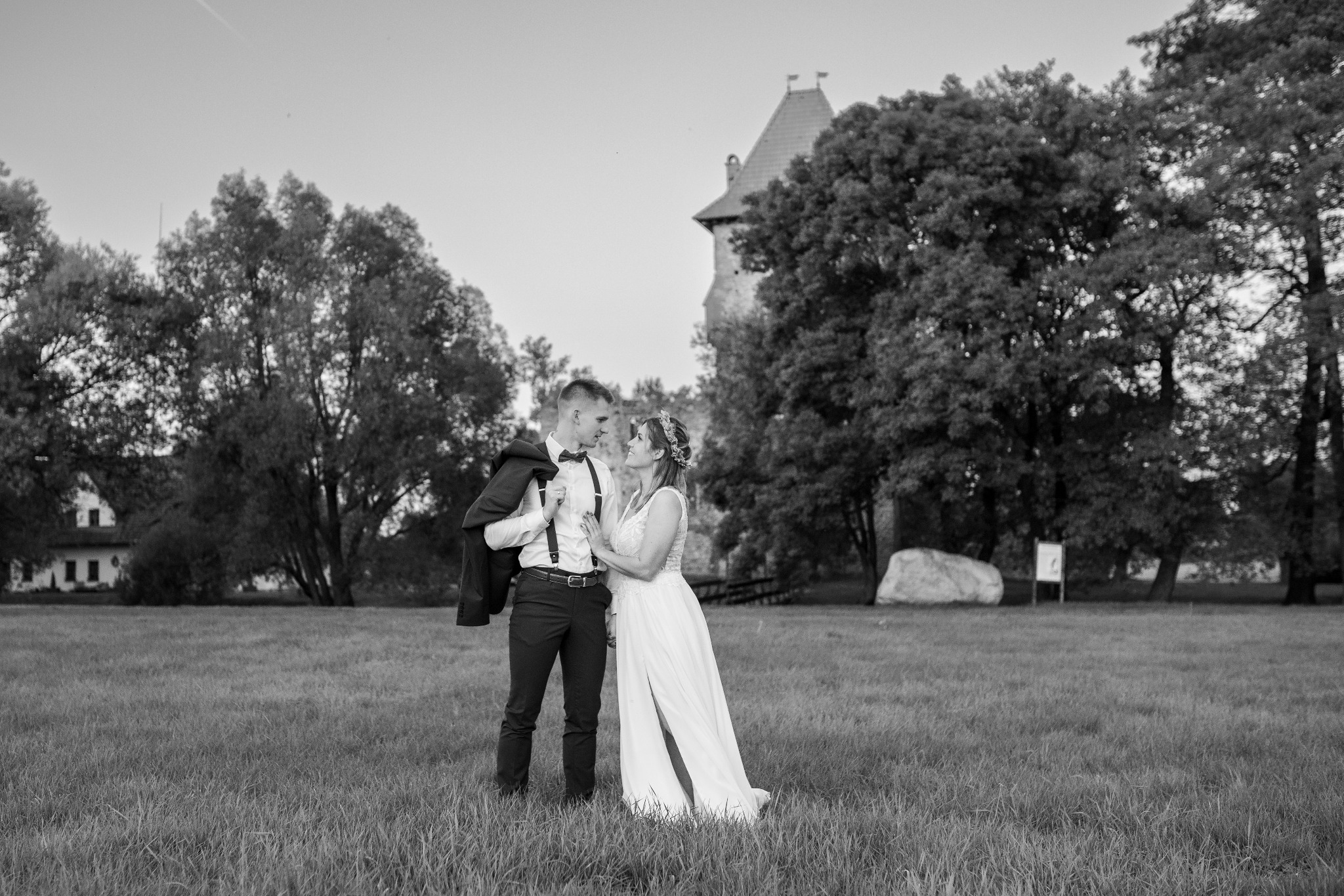 Czarno-białe zdjęcie narzeczonej w koronkowej sukni z rozcięciem i pana młodego z marynarką przewieszoną przez ramię, stojących na trawiastym polu z zamkiem w tle.