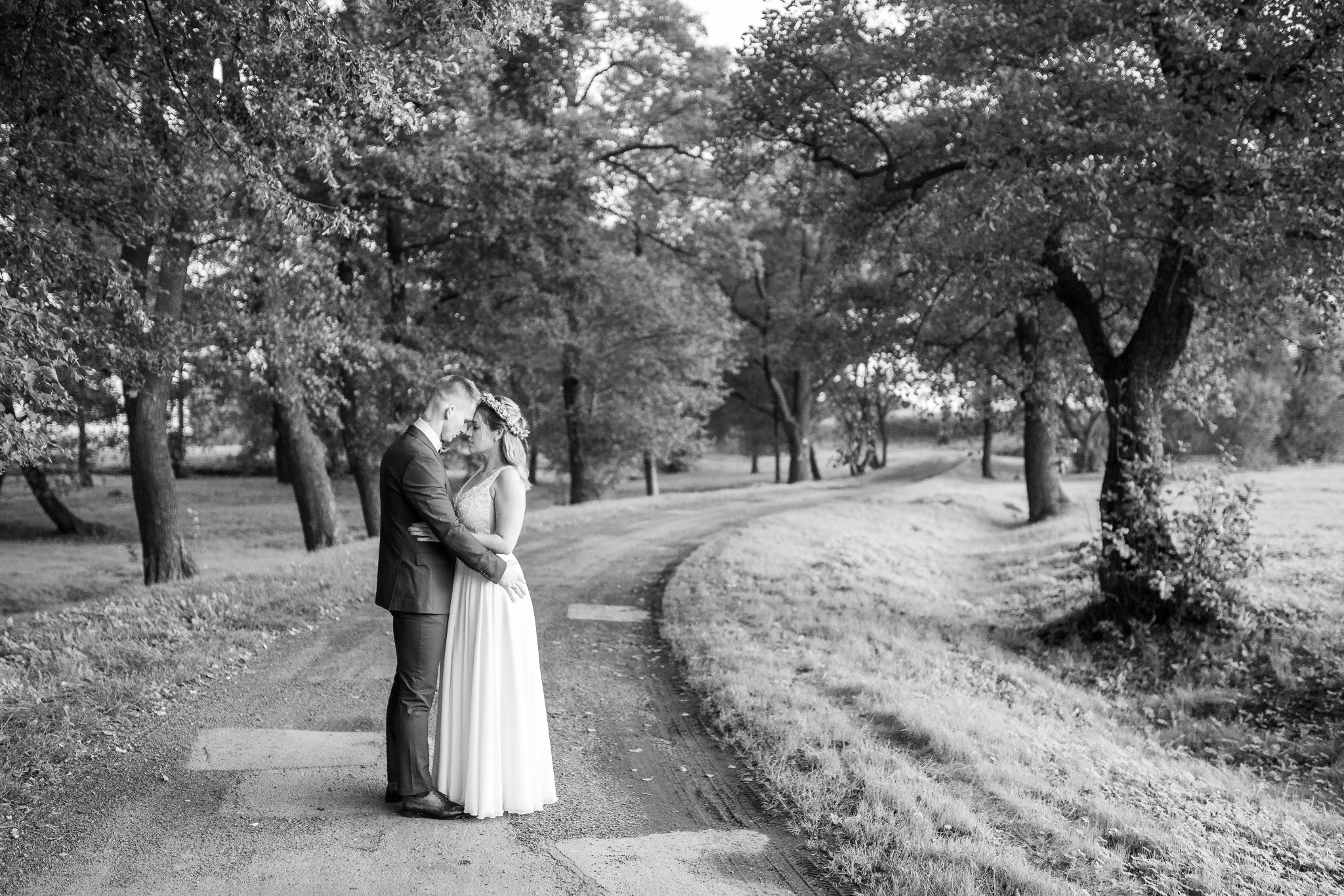 Czarno-białe zdjęcie pary młodej, obejmującej się na leśnej drodze, panna młoda w długiej sukni i wianku, pan młody w garniturze, drzewa w tle.