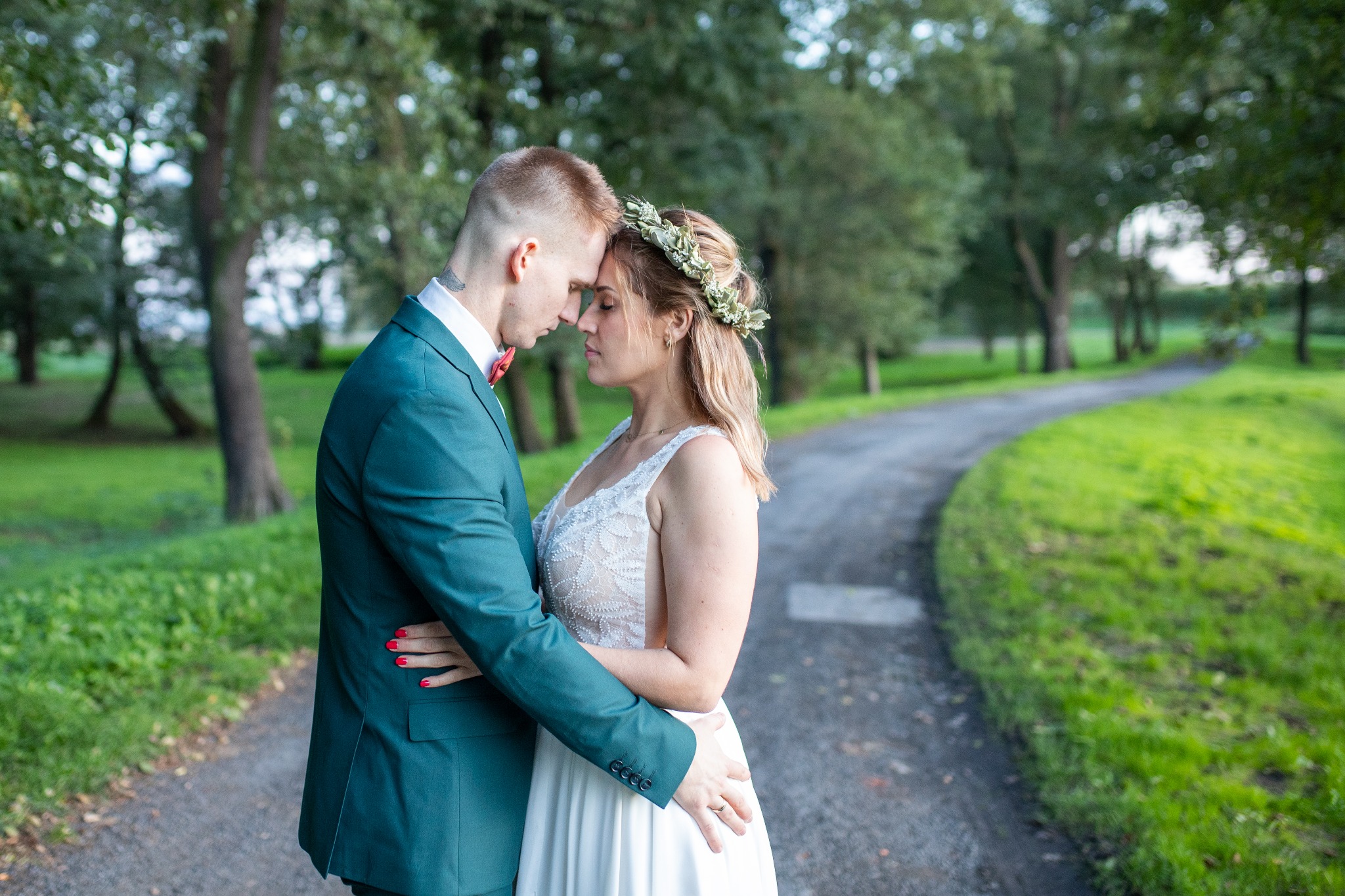 Para młoda w objęciach na leśnej ścieżce: mężczyzna w szmaragdowym garniturze i muszce, kobieta w koronkowej sukni i wianku.