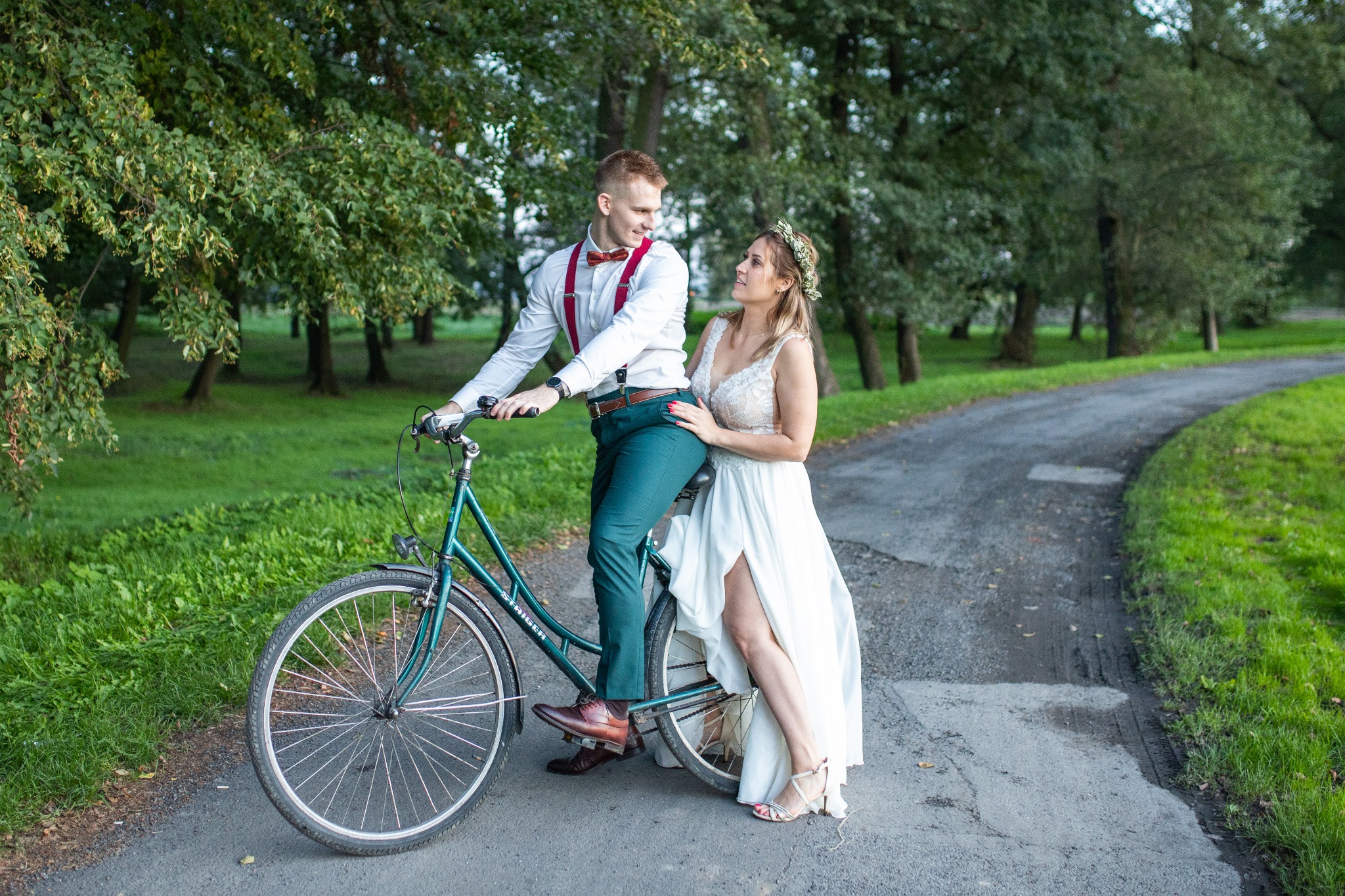 Para młoda w strojach ślubnych pozuje na zielonym rowerze na leśnej ścieżce, panna młoda w wianku opiera się o kierownicę.