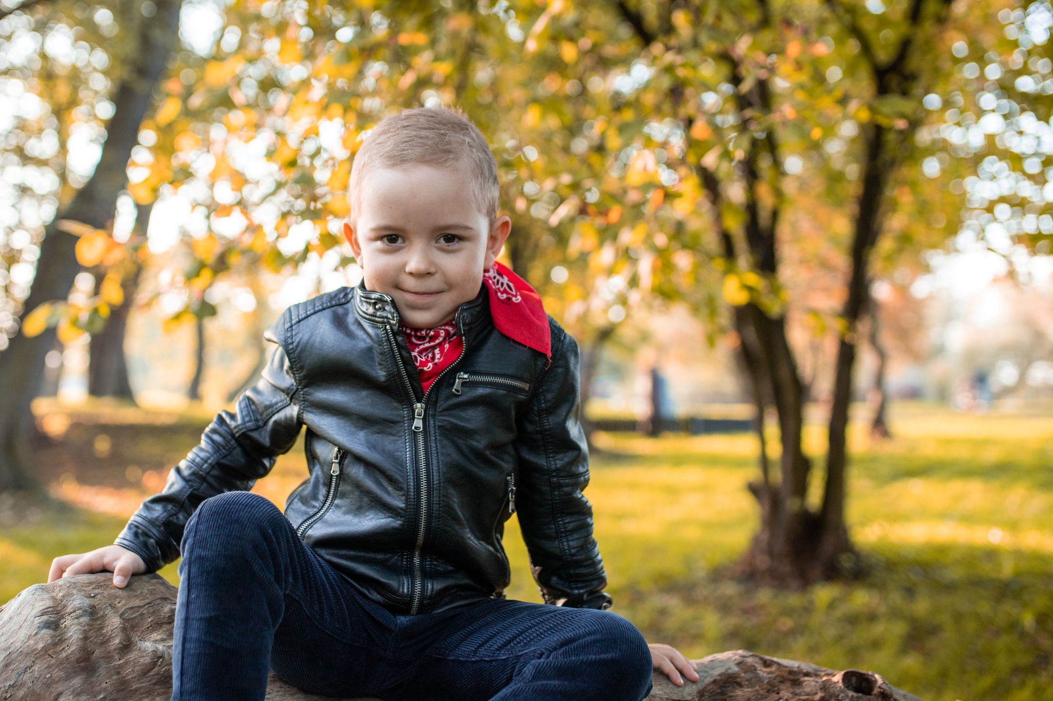 Uśmiechnięty chłopiec w czarnej skórzanej kurtce i czerwonej bandanie siedzi na spróchniałym pniu drzewa w jesiennym parku, z rozmyte drzewa w tle.
