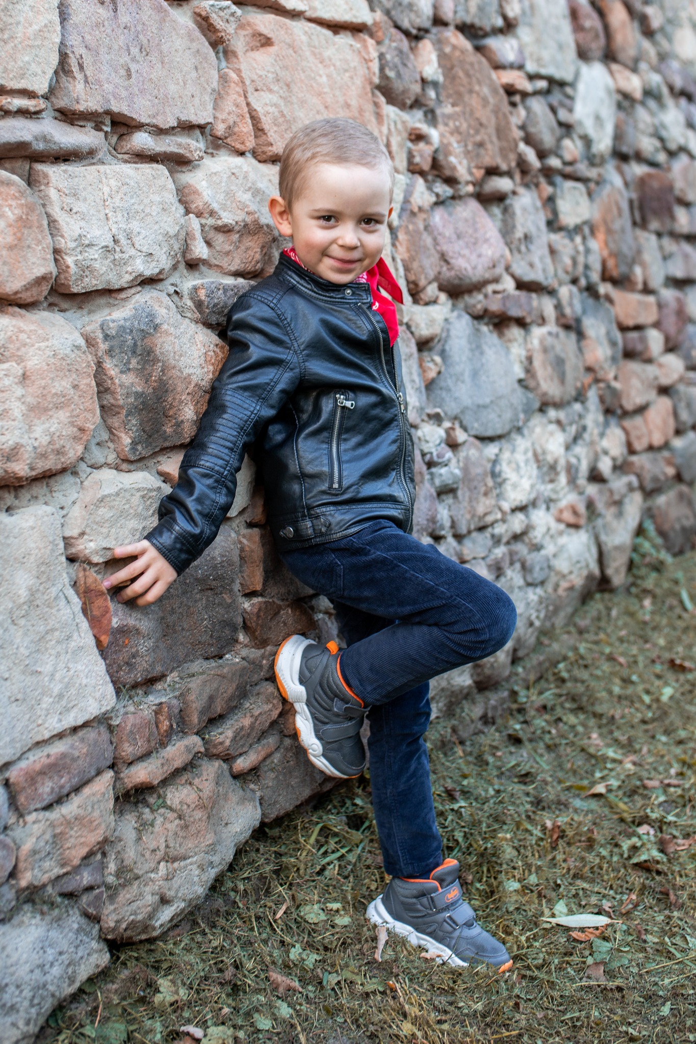 Uśmiechnięty chłopiec w skórzanej kurtce i granatowych spodniach, oparty o kamienną ścianę z czerwoną apaszką.