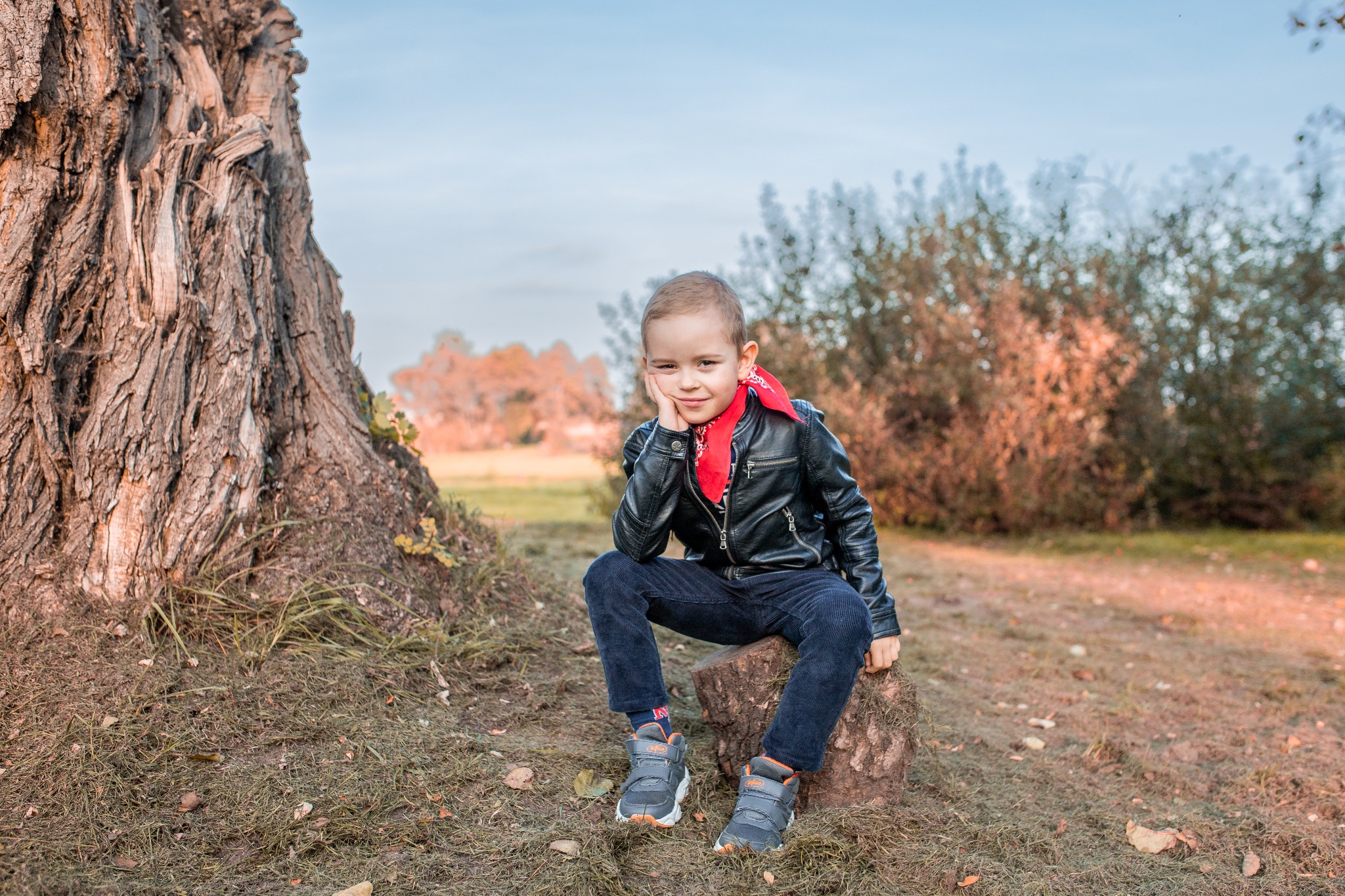 Chłopiec w skórzanej kurtce i czerwonej bandanie siedzi na pniu drzewa, pozując w jesiennej scenerii.