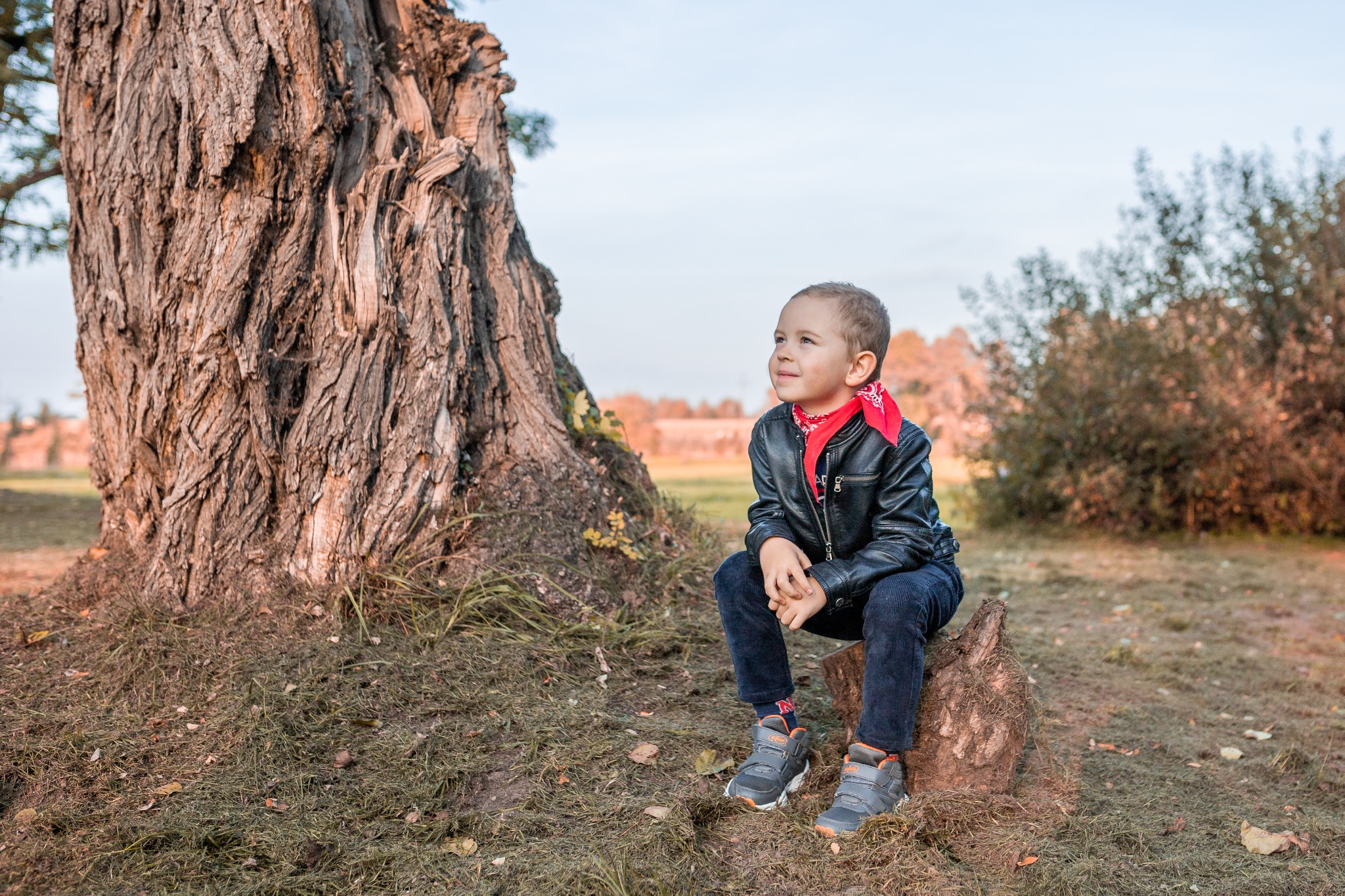 Chłopiec w czarnej skórzanej kurtce i czerwonej bandanie siedzi na pniu drzewa, spoglądając w dal na tle jesiennego krajobrazu.