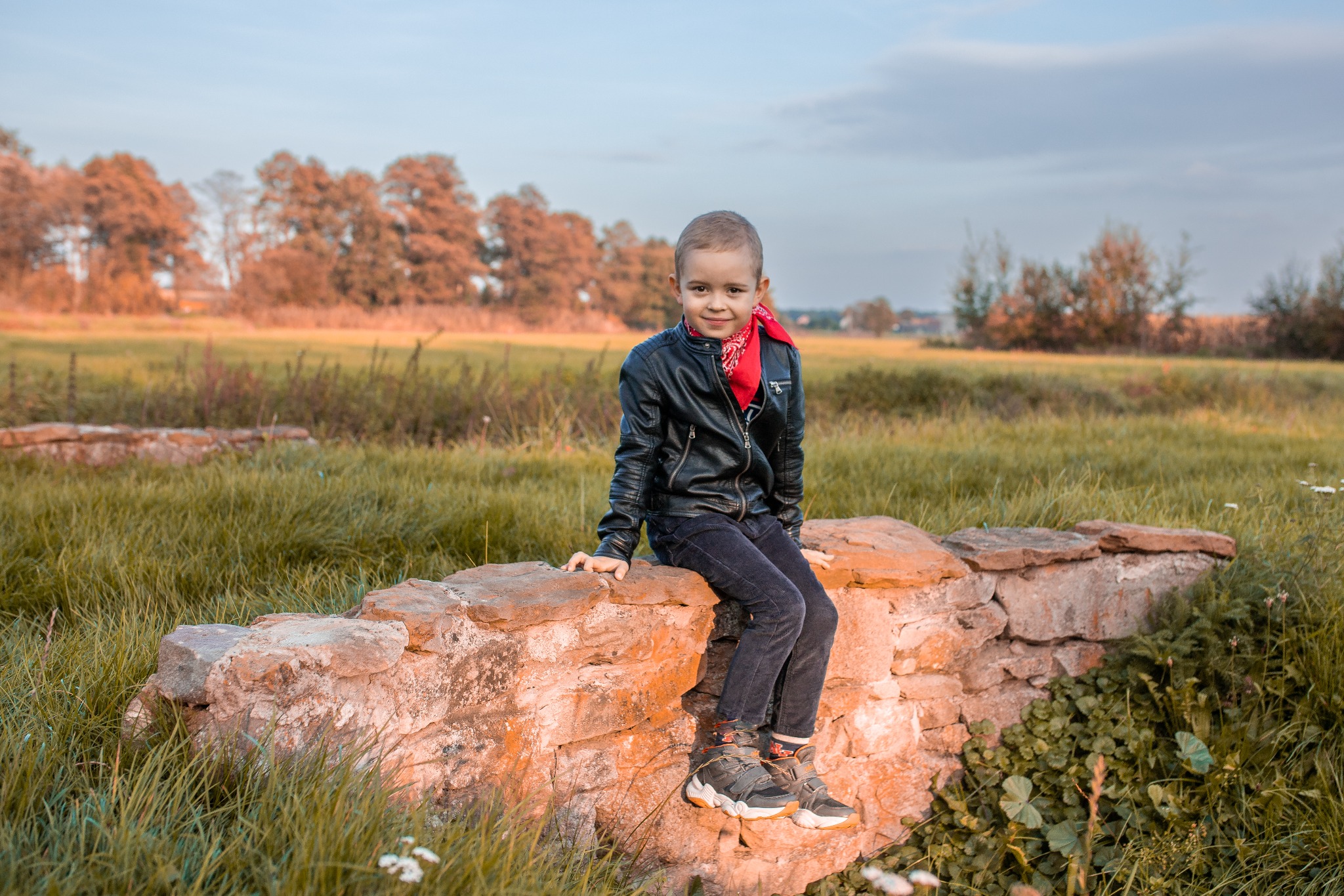 Chłopiec w czarnej skórzanej kurtce i czerwonej bandanie siedzi na niskim kamiennym murku na tle łąki i drzew.