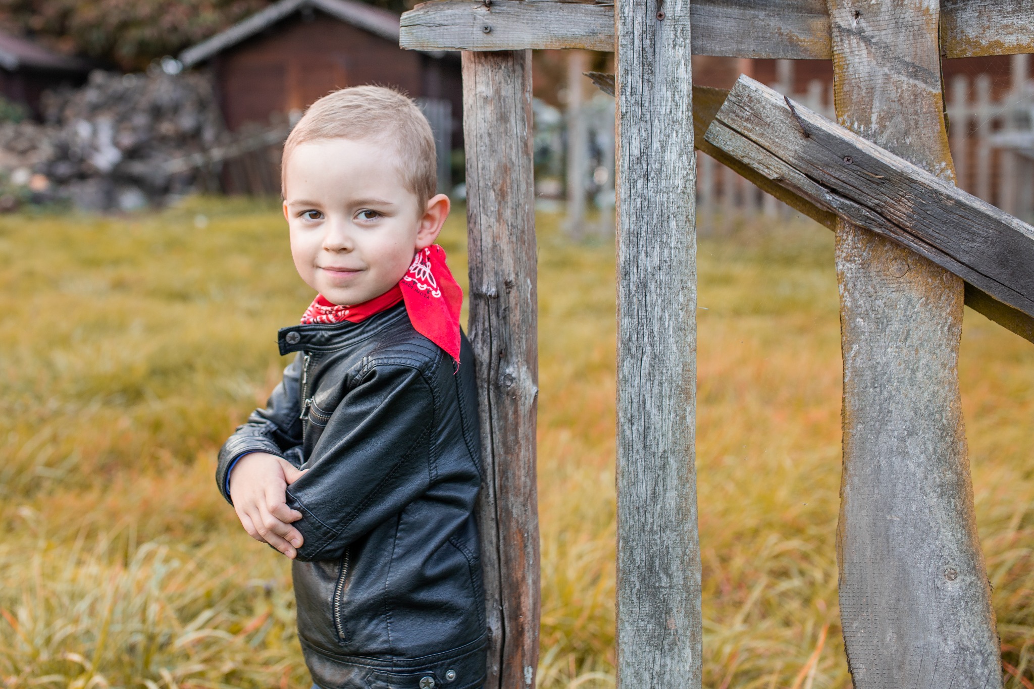 Portret uśmiechniętego chłopca w czarnej skórzanej kurtce i czerwonej bandanie, opierającego się o szary, drewniany płot na tle jesiennej trawy i małego domku.