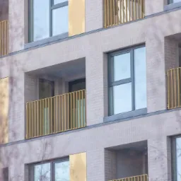 Fragment elewacji budynku z balkonami zabezpieczonymi złotymi, pionowymi barierkami, widok z dołu przez gałęzie drzew