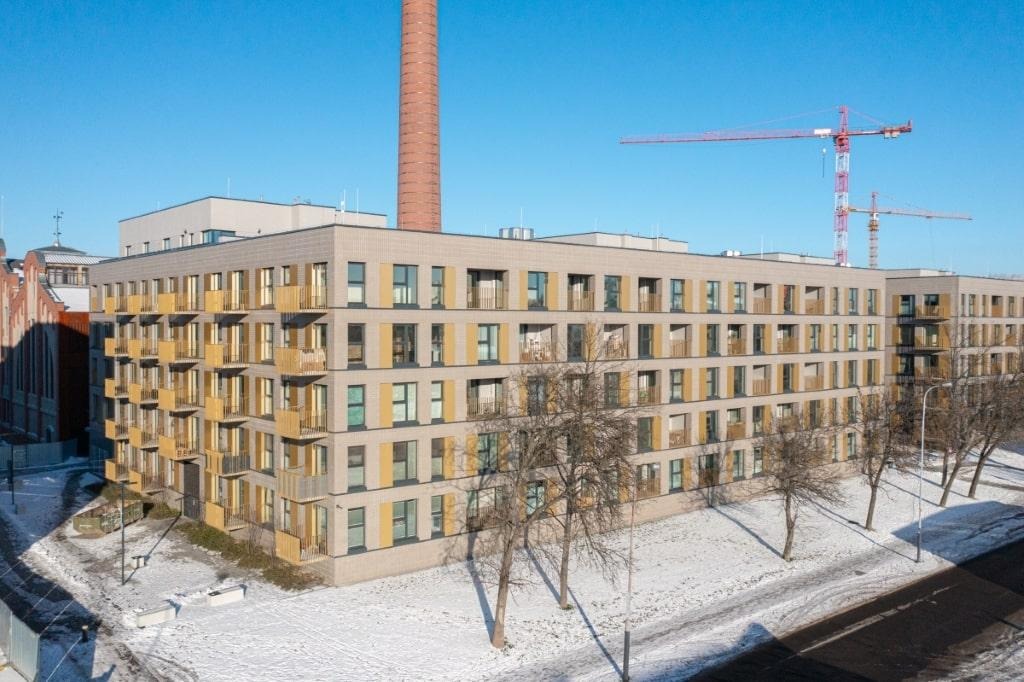 Nowoczesny budynek mieszkalny z żółtymi balustradami balkonowymi, widoczny zimą z perspektywy ulicy, na tle ceglanego komina i żurawi budowlanych.
