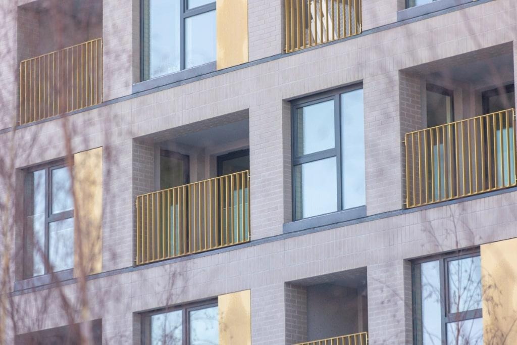 Fragment elewacji budynku z balkonami zabezpieczonymi złotymi, pionowymi barierkami, widok z dołu przez gałęzie drzew